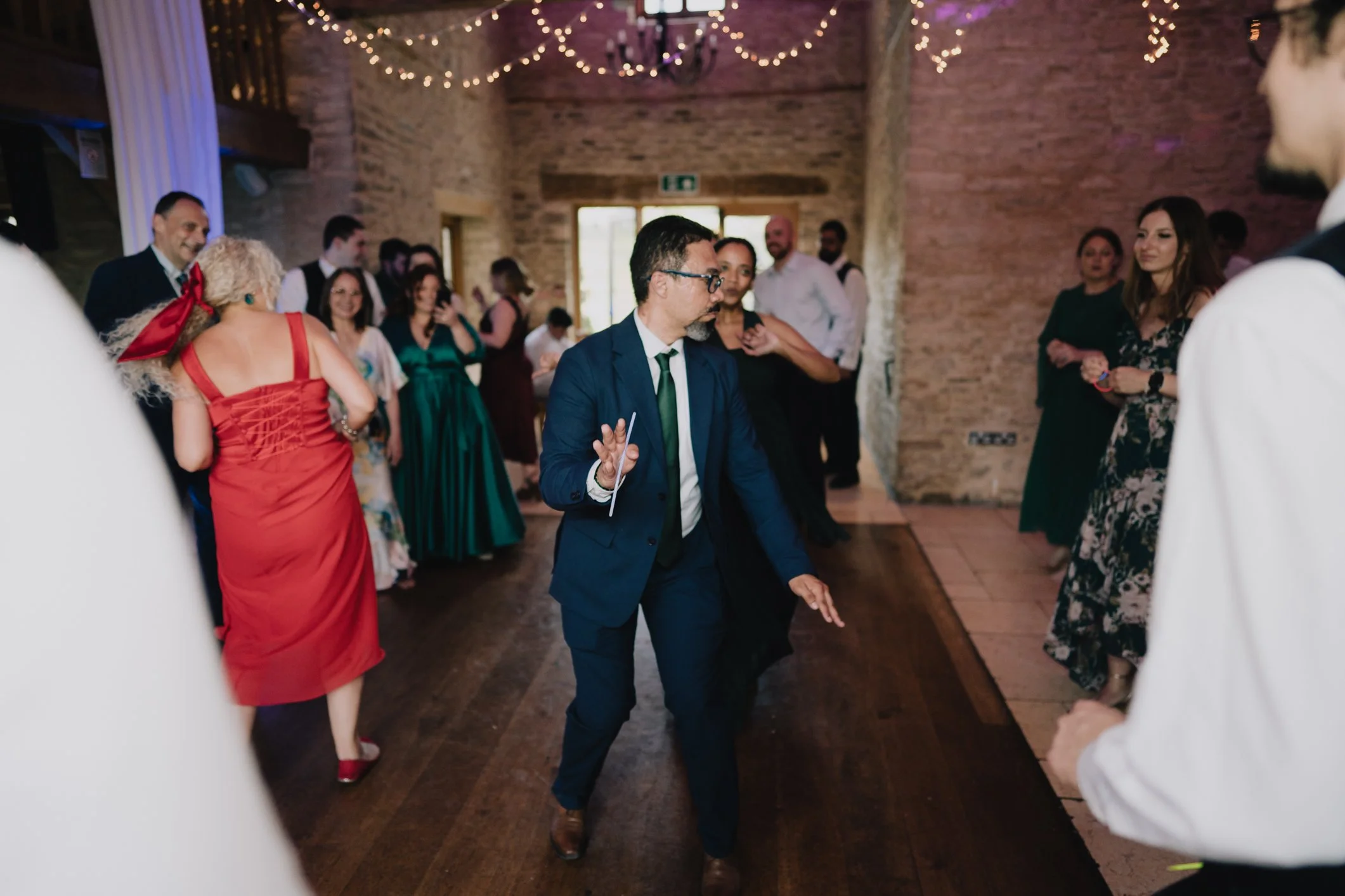 People dancing and socializing at a wedding reception in a rustic venue with string lights