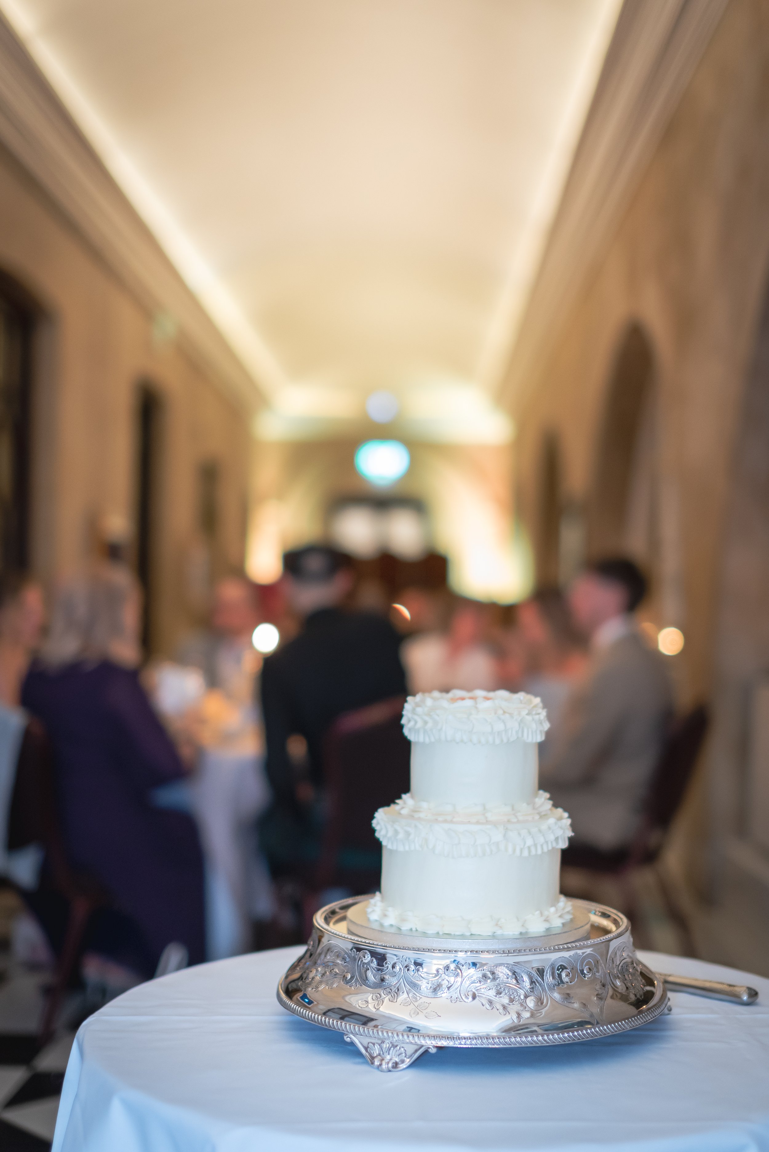 A two-tier white wedding cake with decorative frosting, placed on a silver ornate cake stand on a round table covered with a white tablecloth at a wedding reception. Blurred guests are sitting at tables in an elegant hall in the background.