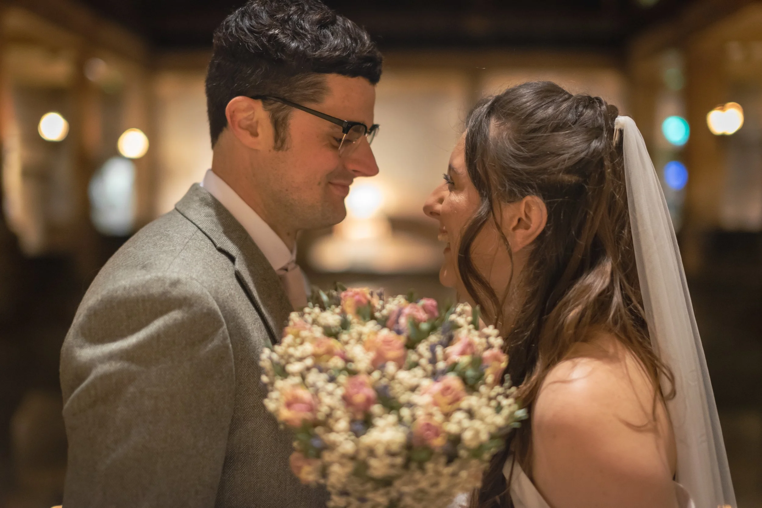 A bride and groom face to face smiling with their noses almost touching, the bride holds a bouquet of pink and white flowers, both are dressed in wedding attire, in a warmly lit indoor setting at Roman Baths.