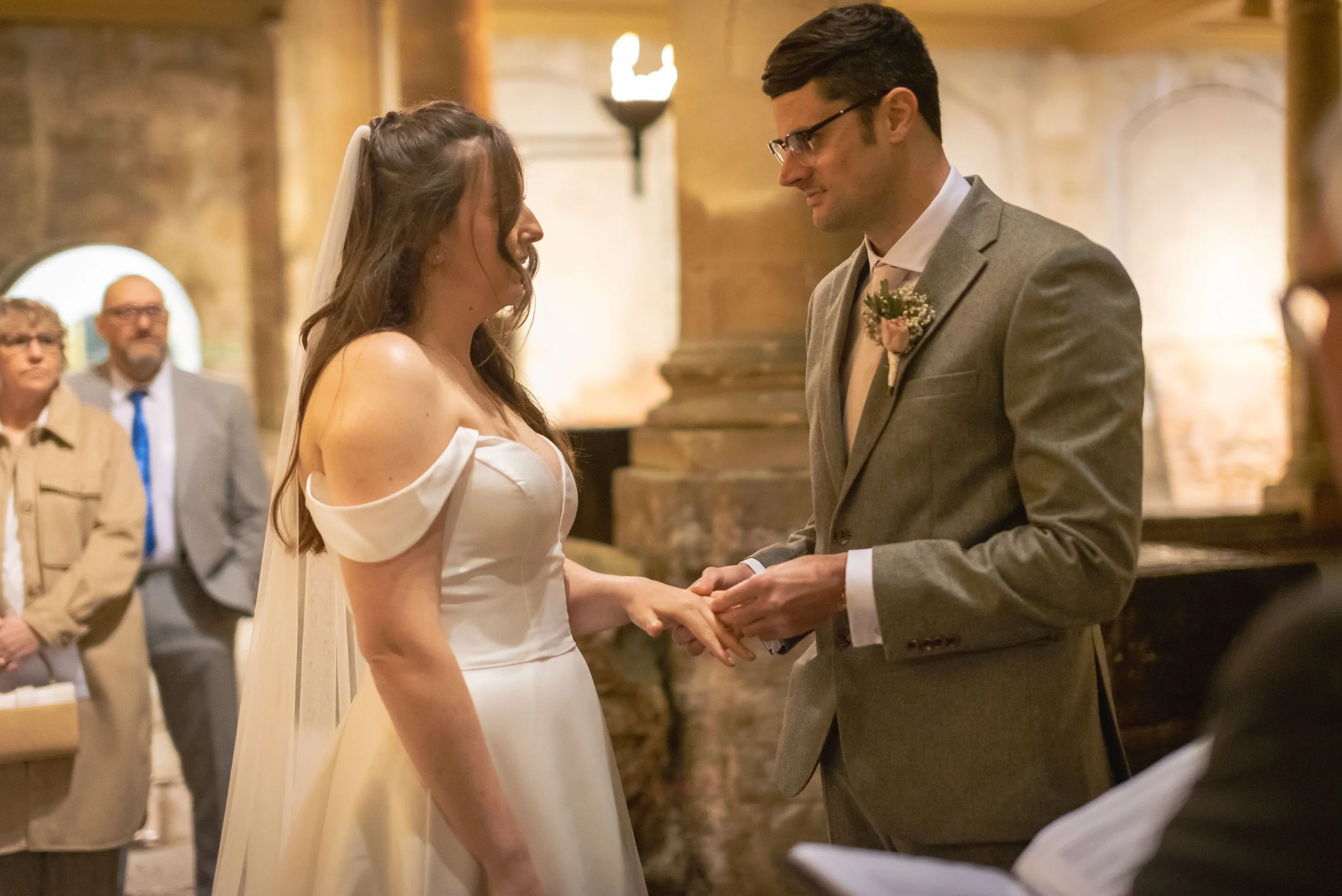 A bride and groom exchanging wedding vows indoors, with several guests in the background, in Roman Baths