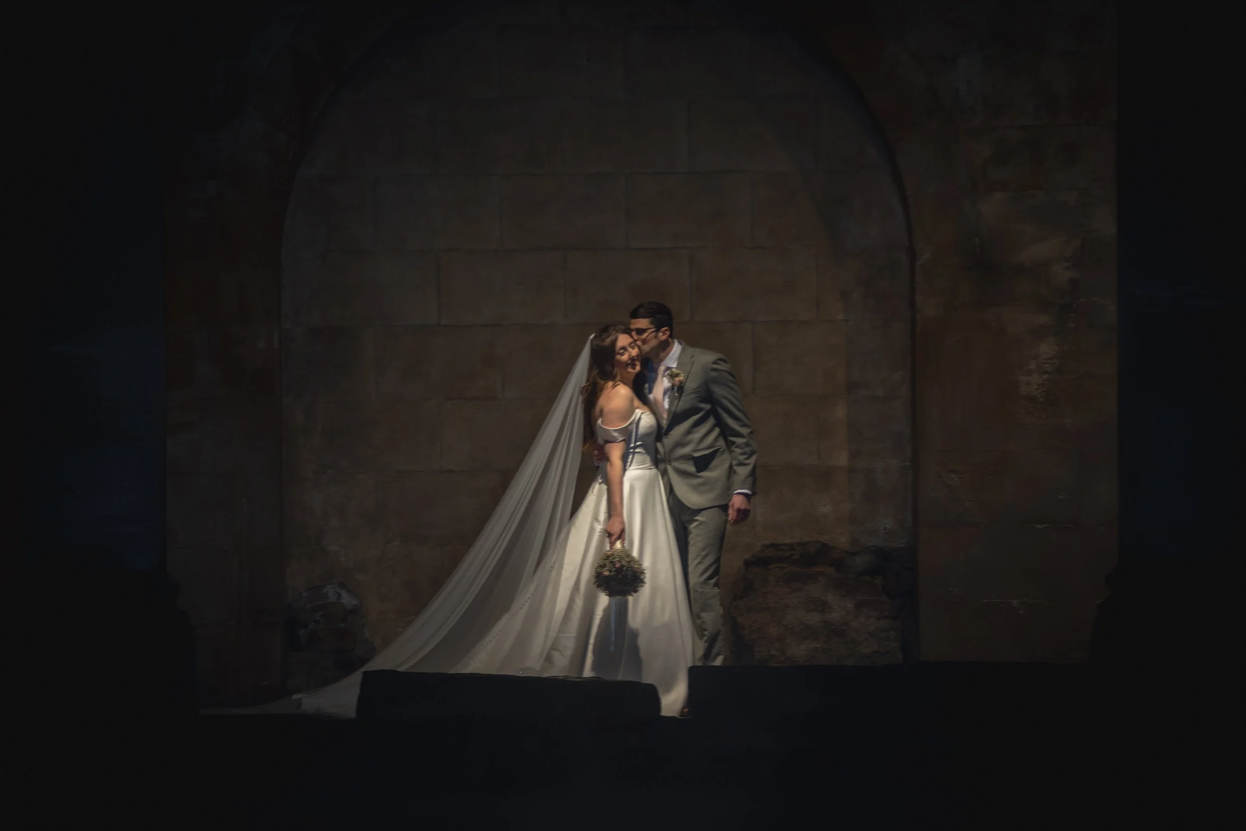 A bride and groom standing close together under an archway, sharing a kiss, with the bride holding a bouquet, in a dimly lit Roman Baths prompted by Wedding Photographer.