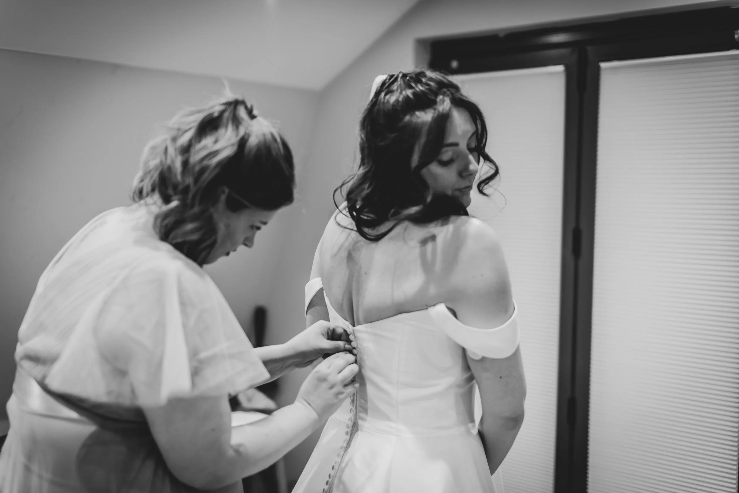 A woman in an off-shoulder wedding dress is being helped by another woman to button up her dress, in a room with sliding doors during bridal prep in Bath.