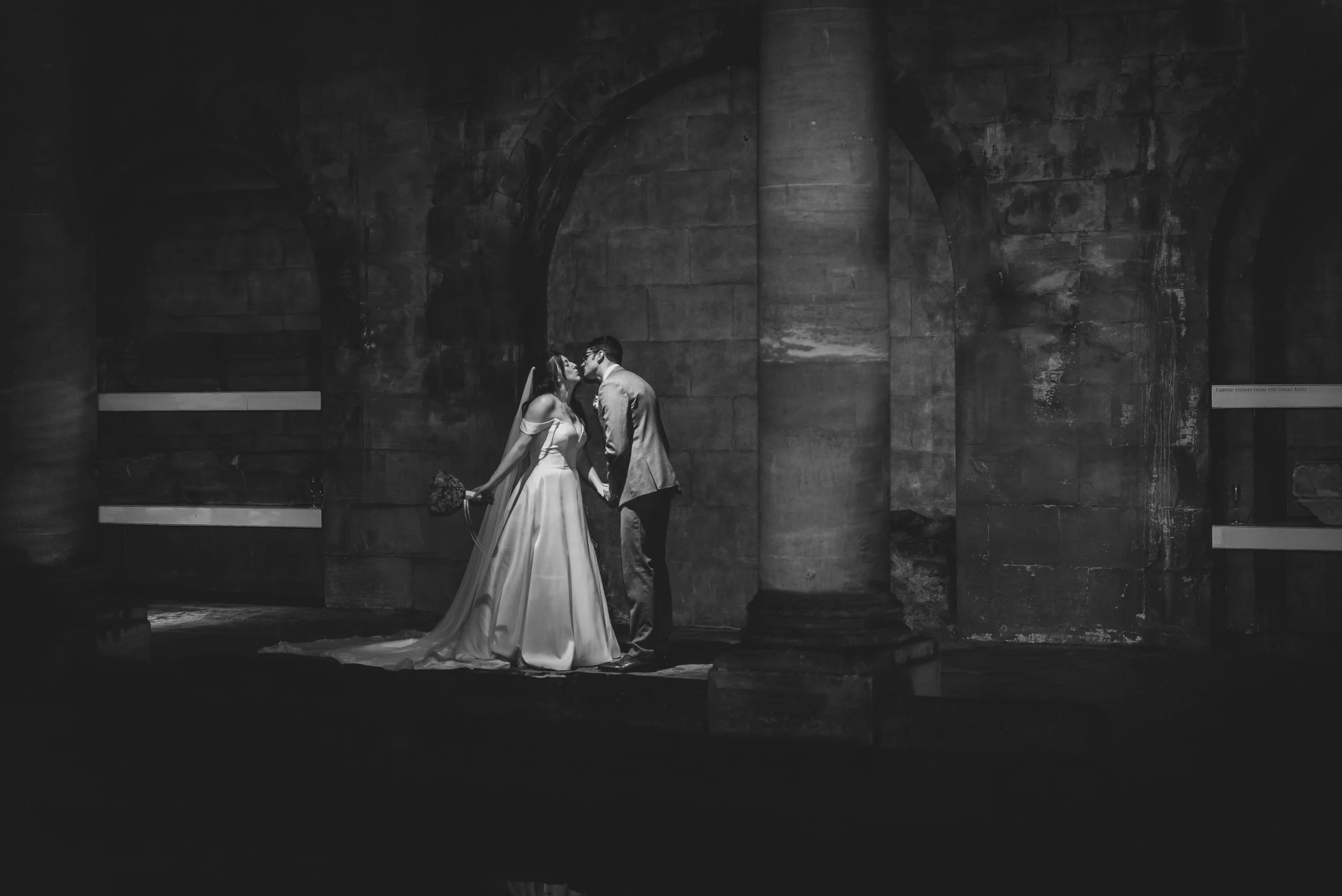 A black and white photo of a bride and groom kissing and holding hands at Roman Bath, Bath.