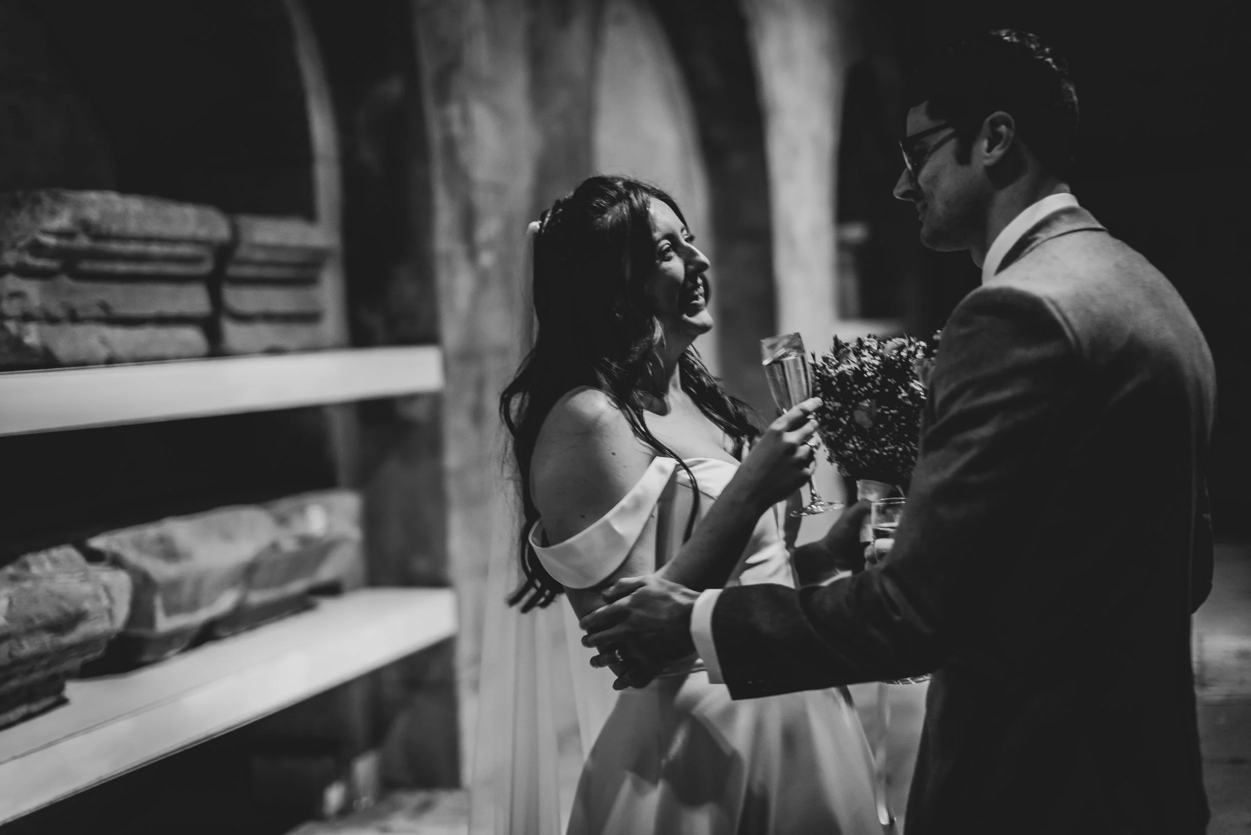 A black and white photograph of a woman and a man at a wedding or celebration. The woman is smiling and holding a glass of champagne, wearing an off-shoulder dress. The man is dressed in a suit and glasses, holding a bouquet of flowers, facing the woman, in an indoor setting with a stone wall in the background.