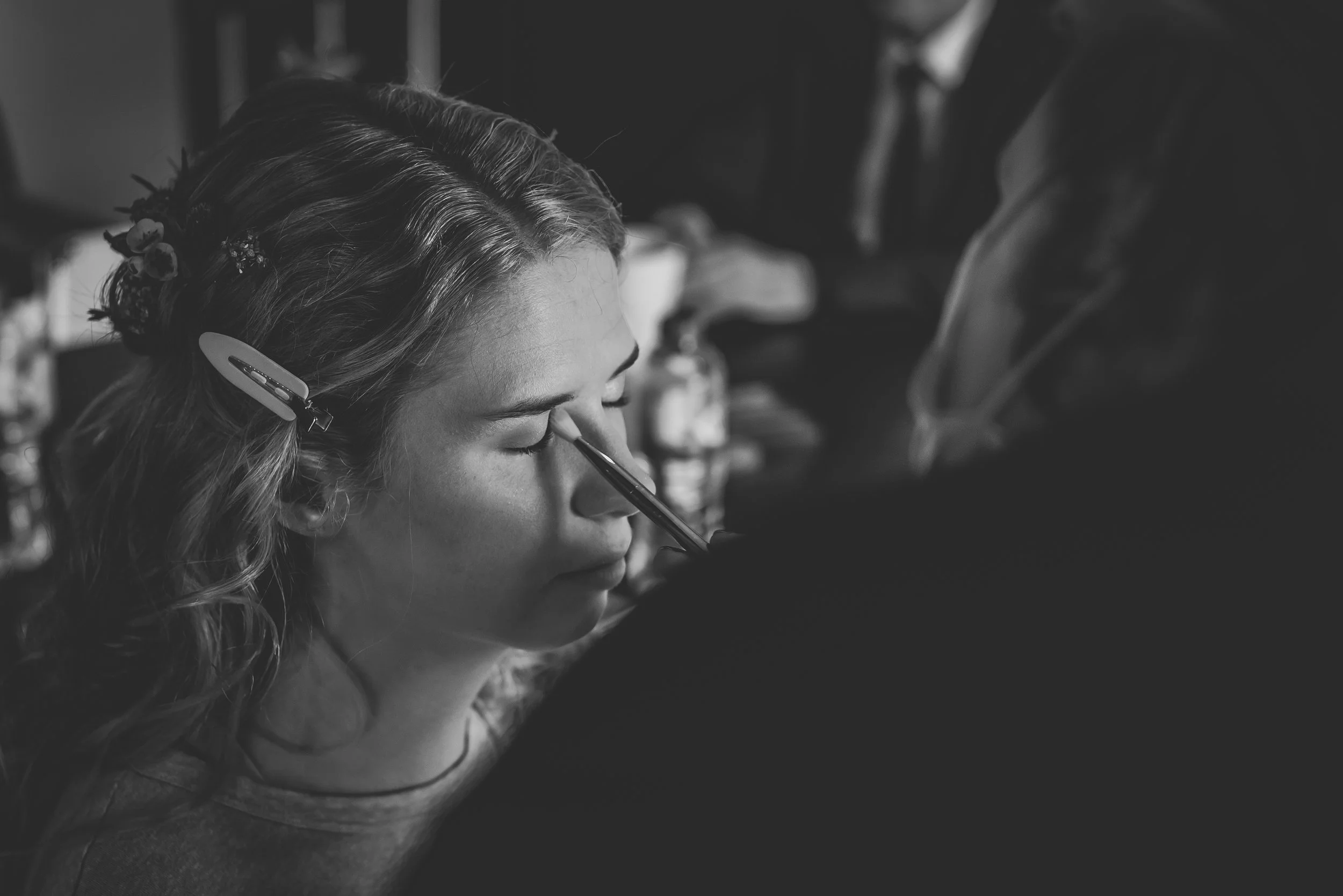 A woman with wavy hair and a floral hairpiece has her eye closed as someone applies makeup with a brush.