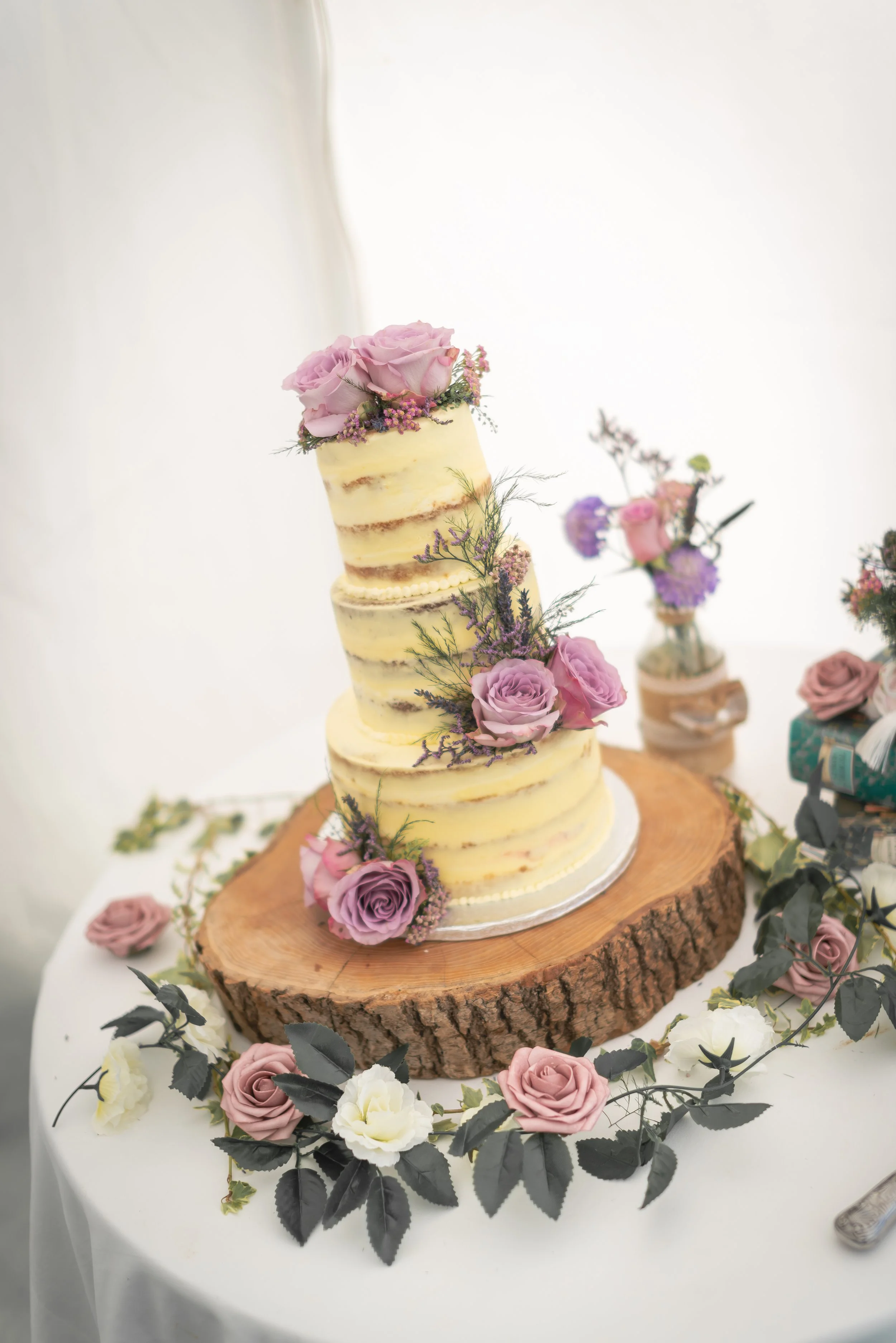 A three-tier naked wedding cake adorned with pink and purple roses and purple accents, placed on a wooden slab, surrounded by pink, white, and purple roses and greenery on a white table.