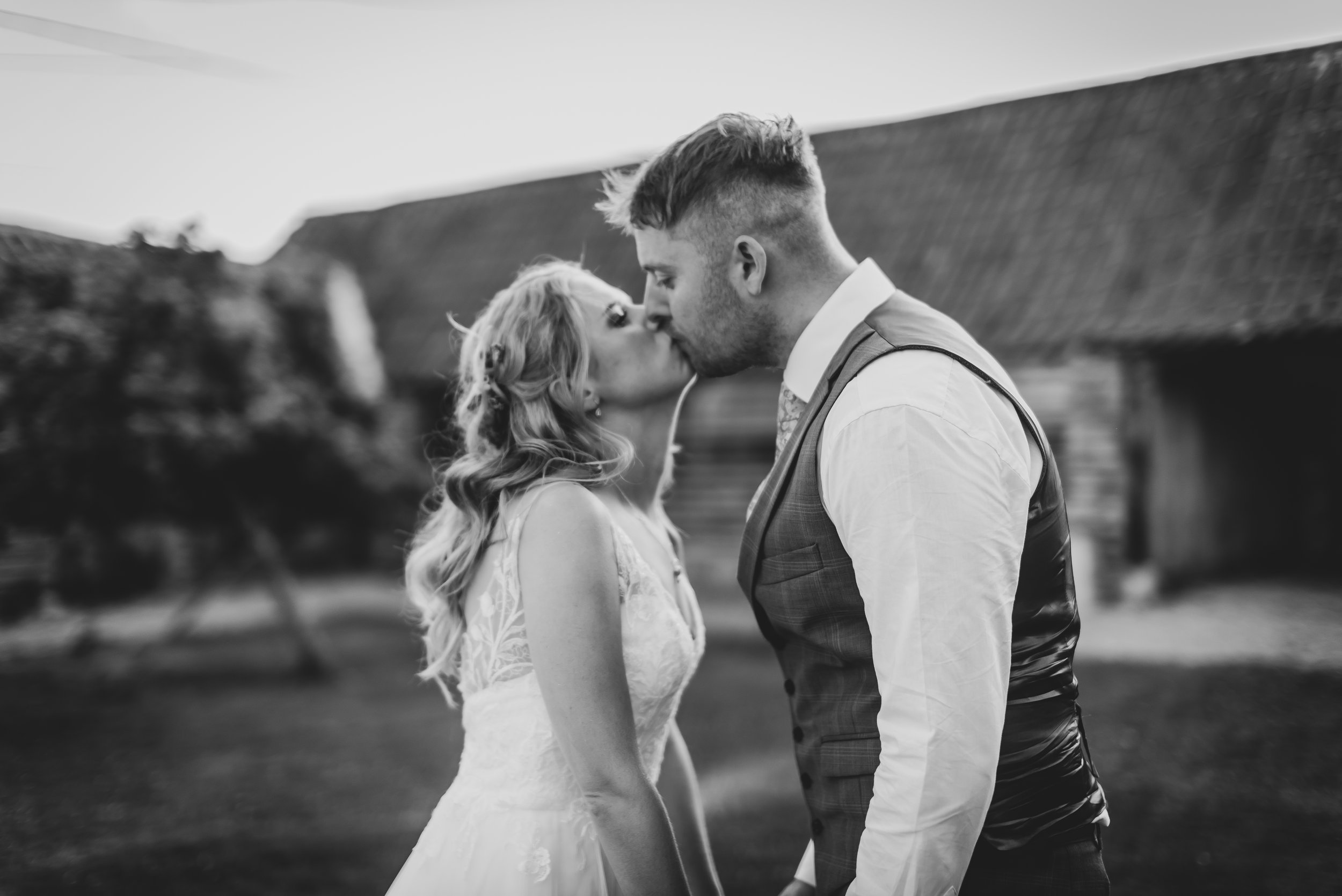 Black and white photo of a couple kissing outdoors, with the woman in a wedding dress and the man in a vest and shirt, near a rustic barn captured by Standerwick Wedding Photographer