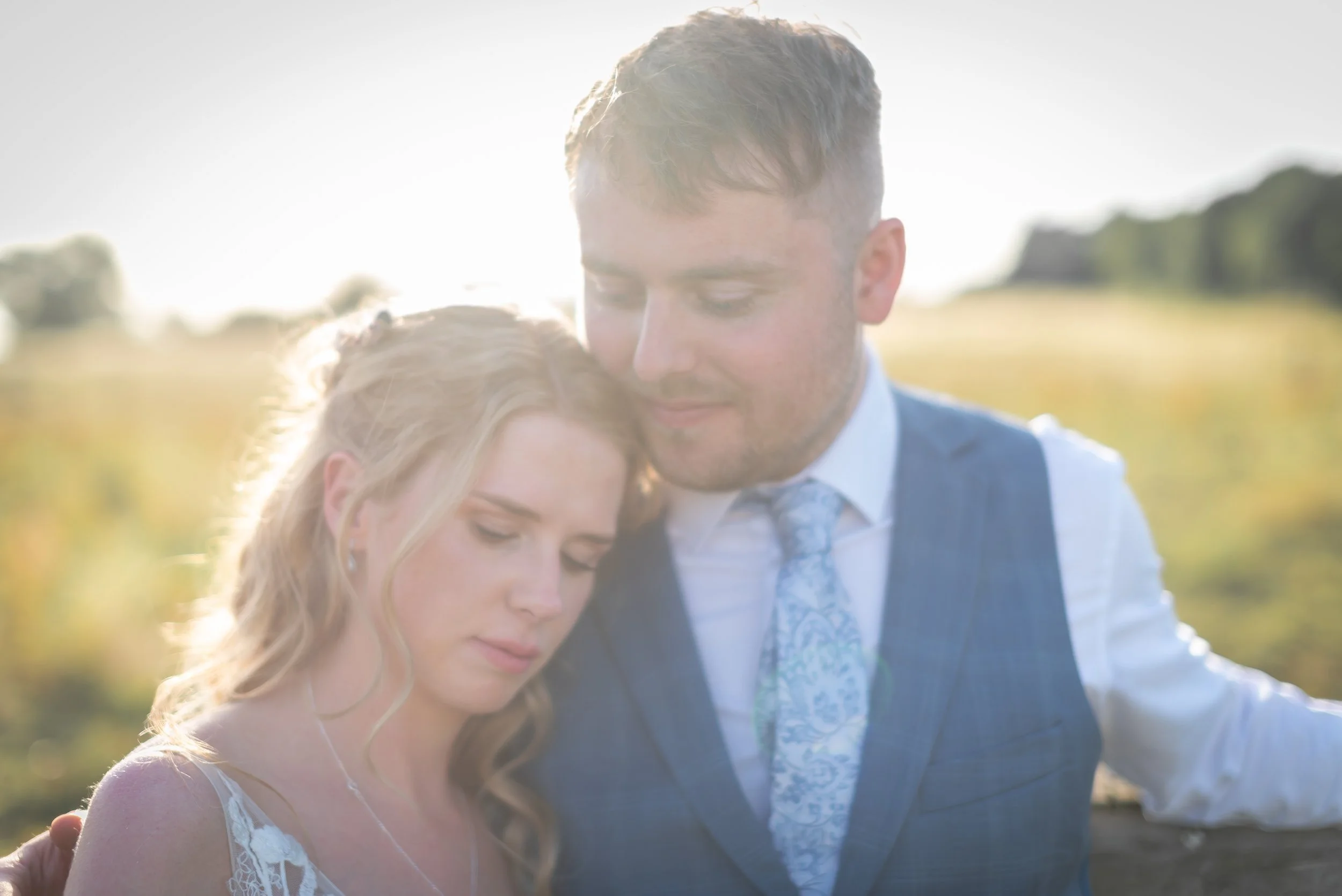 A bride and groom in formal attire, with the woman in a lace dress and the man in a blue suit and floral tie, sharing an intimate moment outdoors with sunlight in the background.