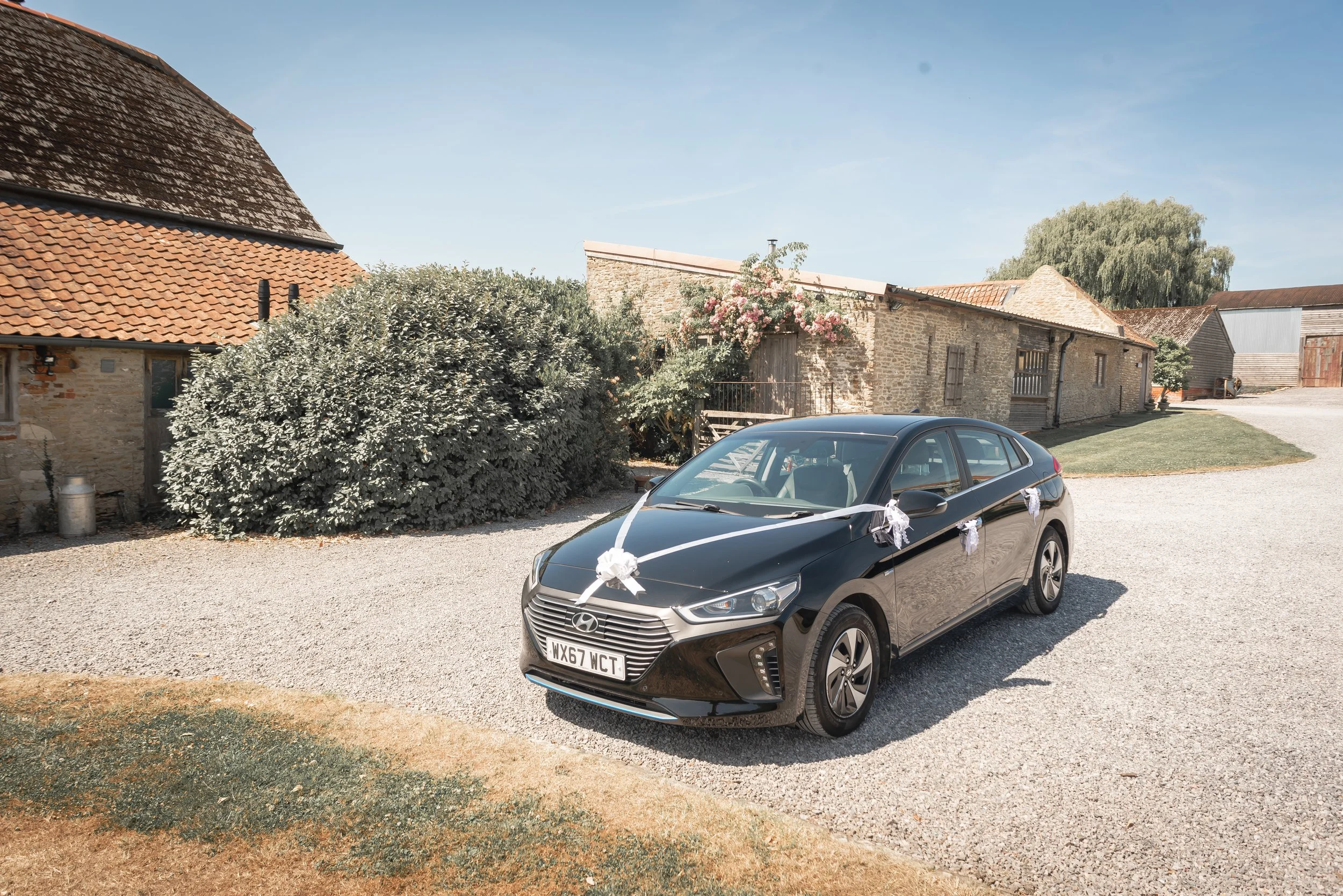A black wedding car decorated with white ribbons and bows, parked outdoors on a gravel driveway in front of rustic buildings and greenery, likely prepared for a wedding or celebration at Court Farm Standerwick