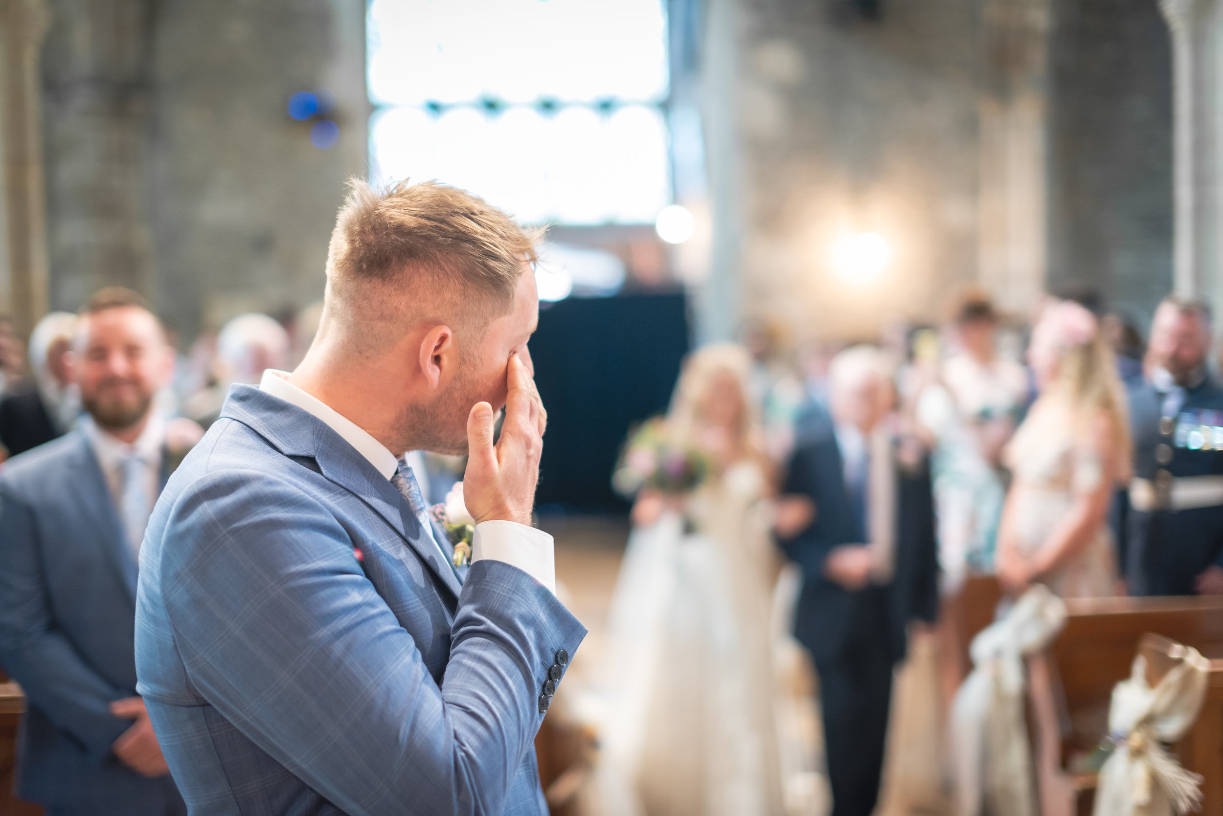A man in a light blue suit is wiping his tear away during a wedding ceremony, with a blurred group of guests behind him in a rustic church setting.