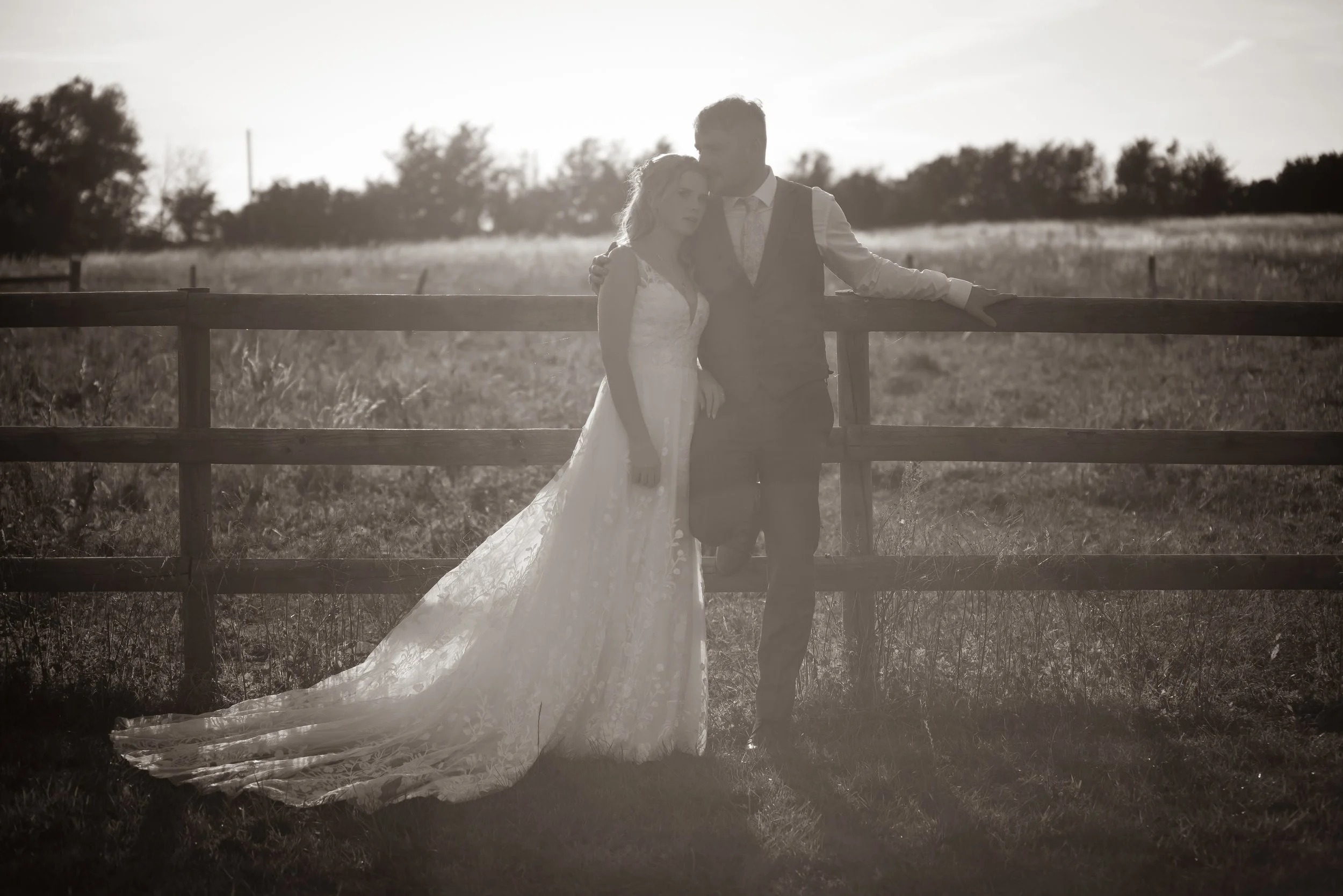 Black-and-white photo of a bride and groom standing together outdoors near a wooden fence at sunset, with trees in the background captured by Standerwick Wedding Photographer at Court Farm.