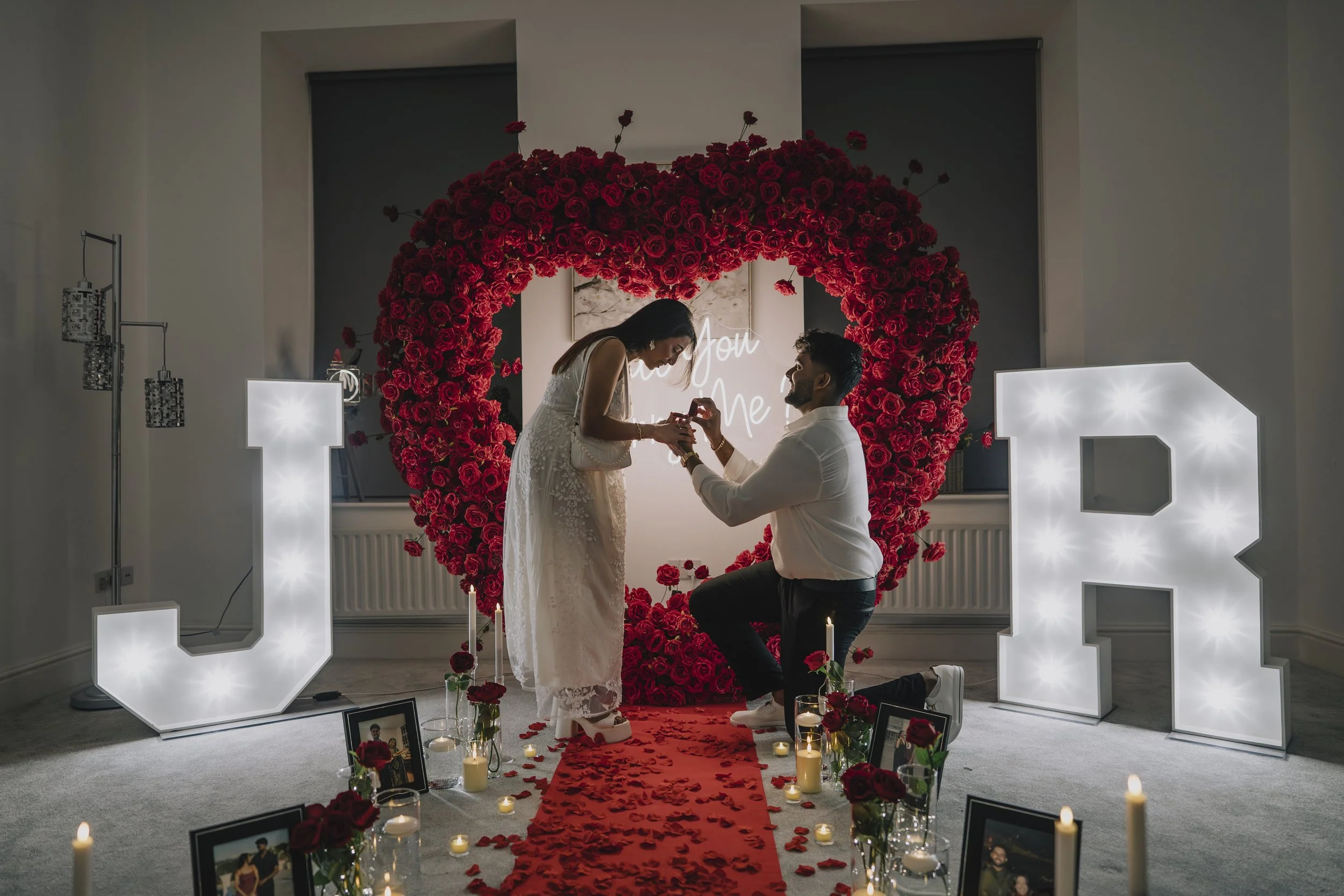 A couple proposes during a wedding ceremony with a heart-shaped floral backdrop, large illuminated letters 'J' and 'R', photo frames, candles, and rose petals on the floor in Bristol.