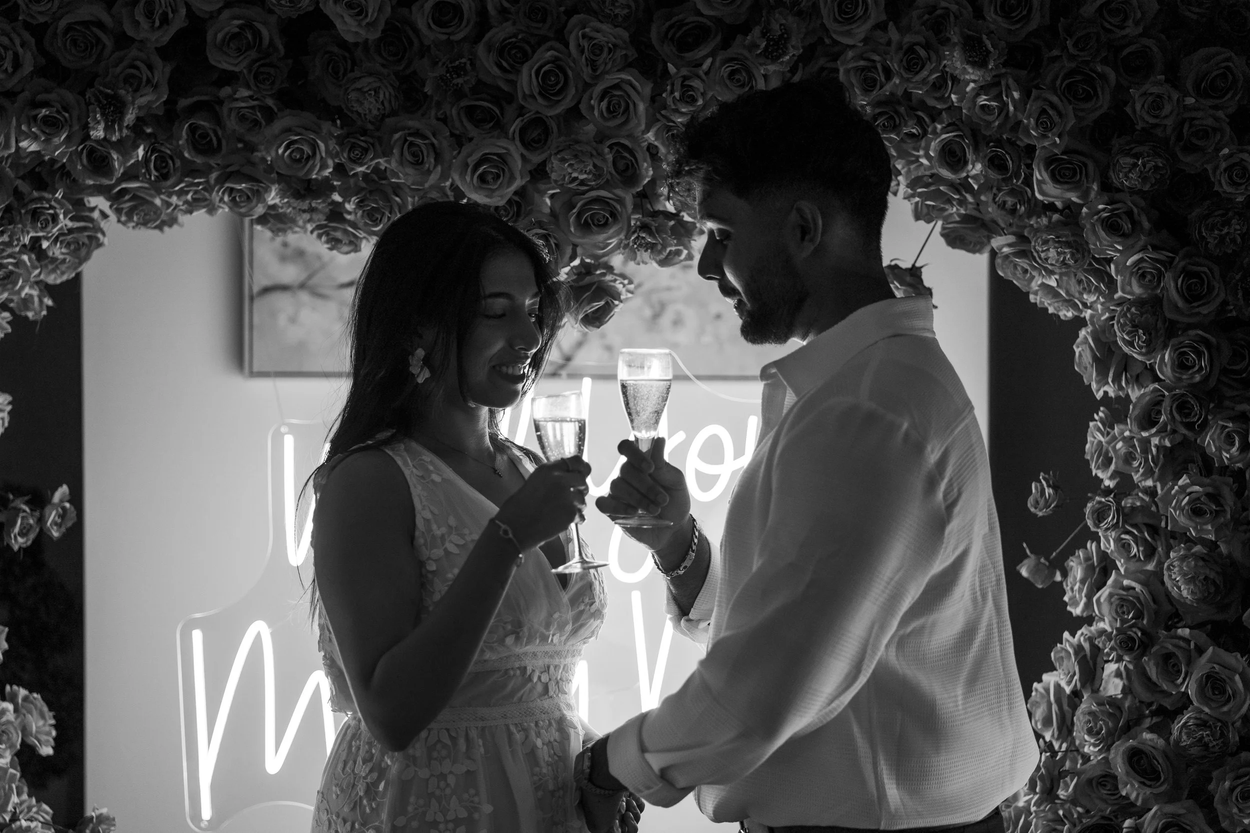 A couple on a date, smiling and holding glasses of champagne, in front of a floral backdrop with a neon sign and flowers after a proposal in Bristol.