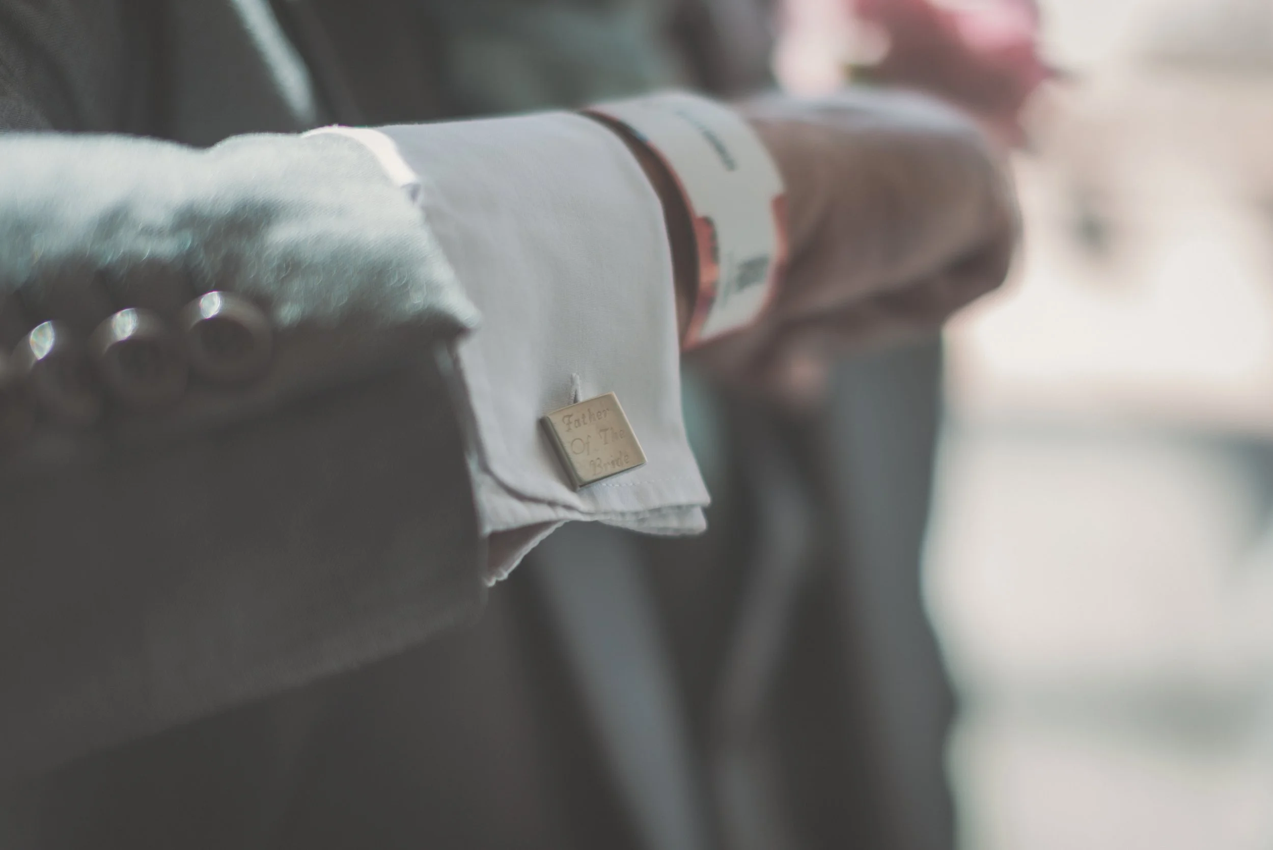 Close-up of a groom's sleeve with a white shirt cuff and a small metal pin that reads "Father Of The Bride", with a blurred guest's arm wearing a hospital wristband in the background.