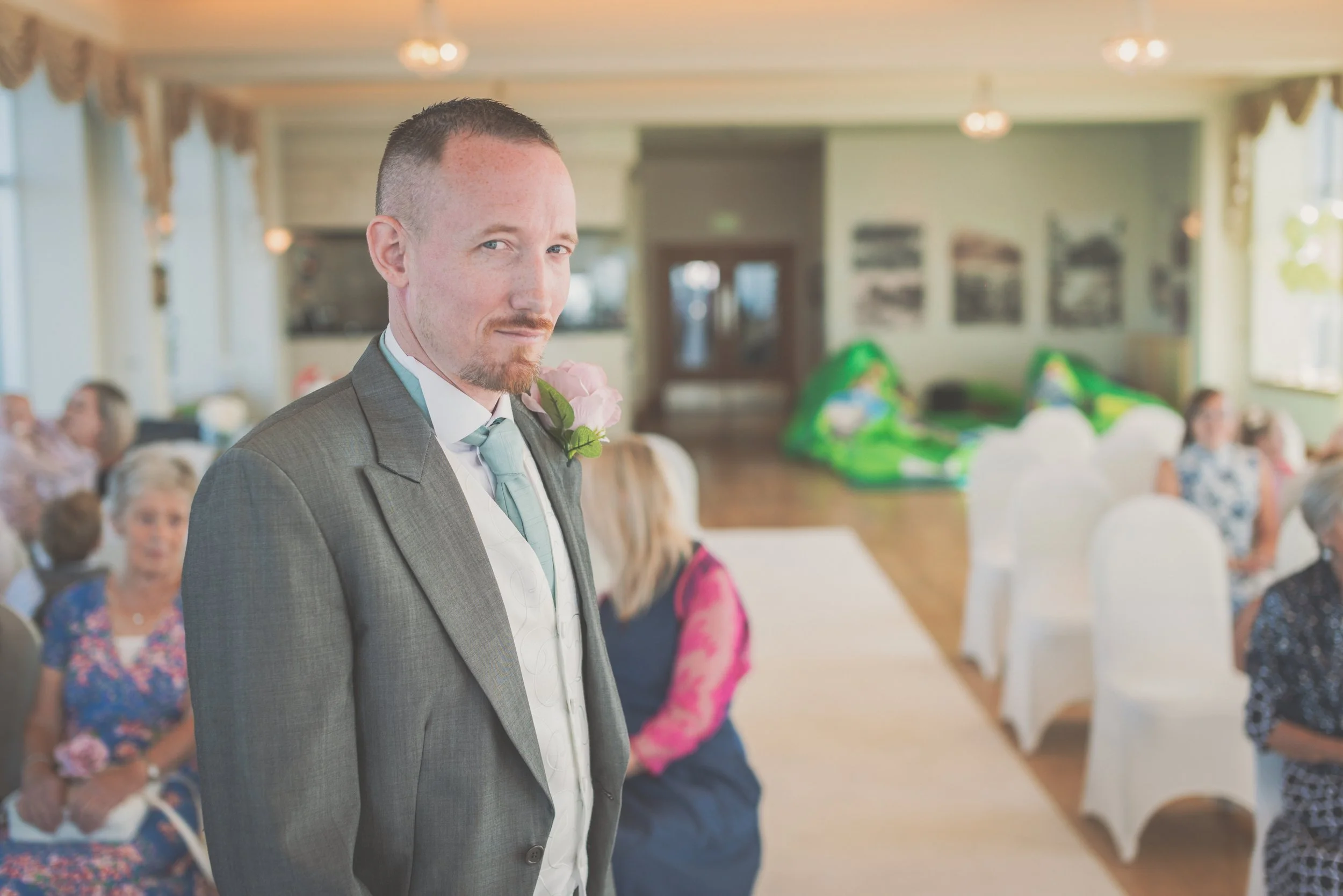 A man in a gray suit with a boutonniere stands in a decorated indoor venue, likely a wedding reception, with guests seated in the background.