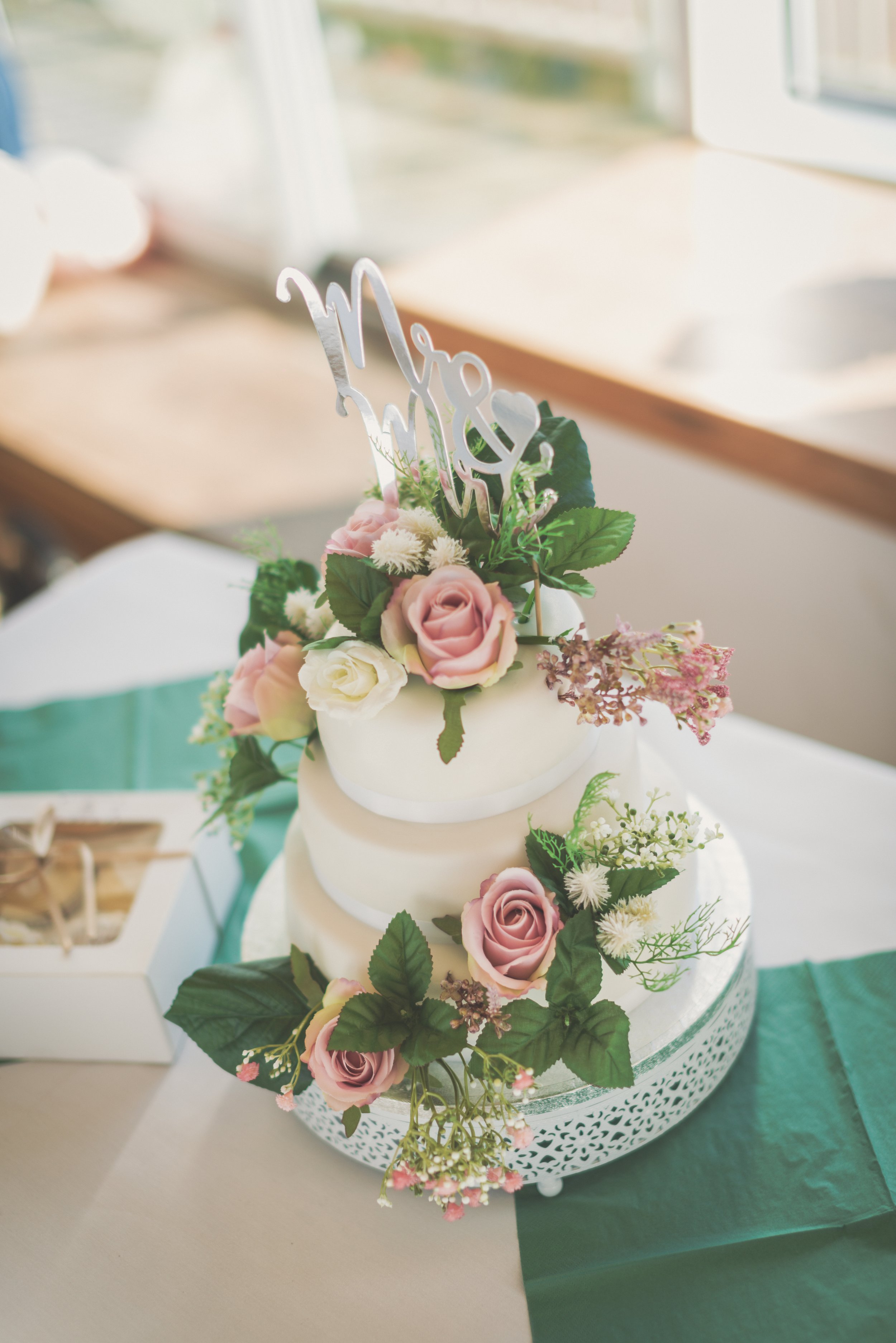 A white tiered wedding cake decorated with pink roses, white flowers, green leaves, and a silver 'Mr & Mrs' cake topper, placed on a decorative white cake stand with a green and beige tablecloth underneath.