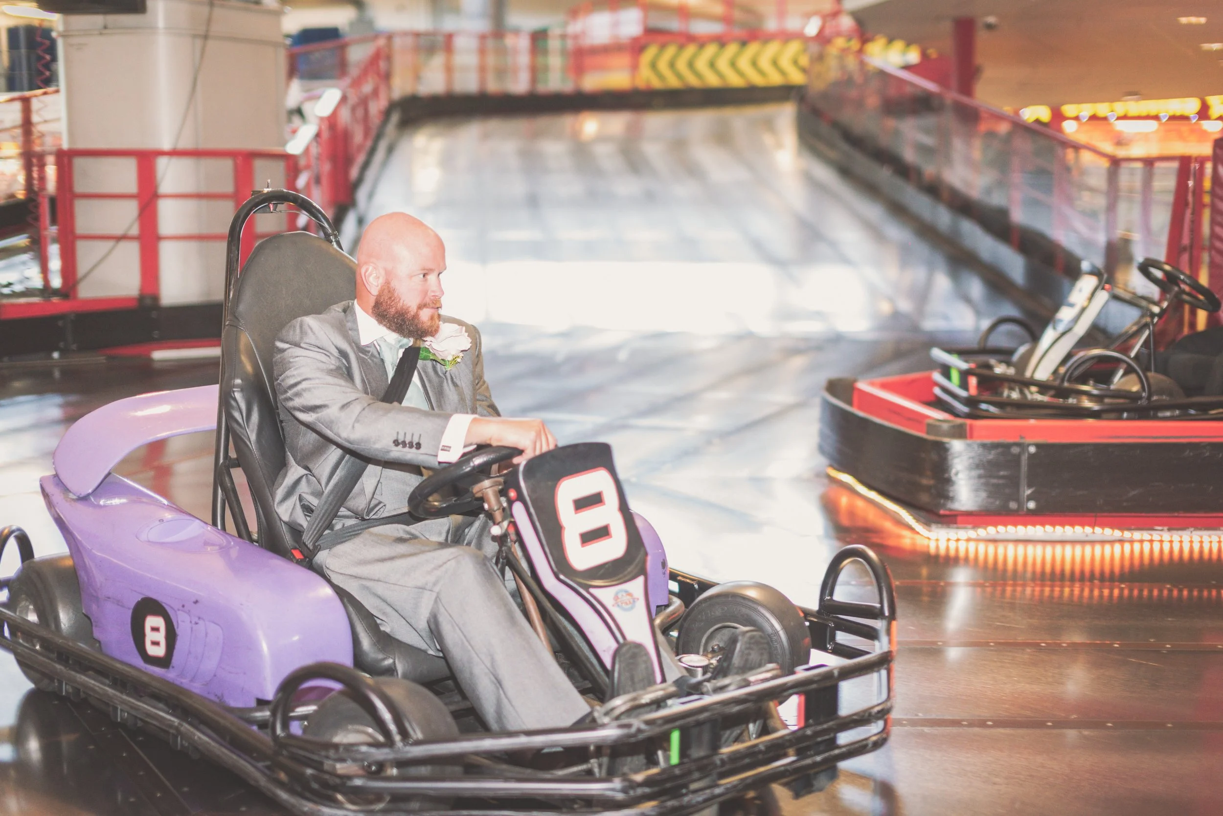 A man in a gray suit and white shirt rides a purple and black indoor go-kart on a race track, with a focused expression at the Grand Pier, Weston-super-Mare.