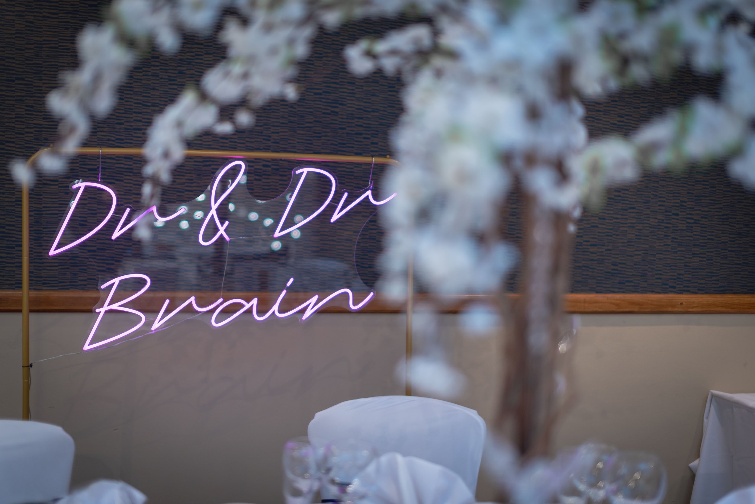 Neon sign with pink and purple words 'Dr & Dr Brain' in a room decorated for an event, with blurred white flowers in the foreground.