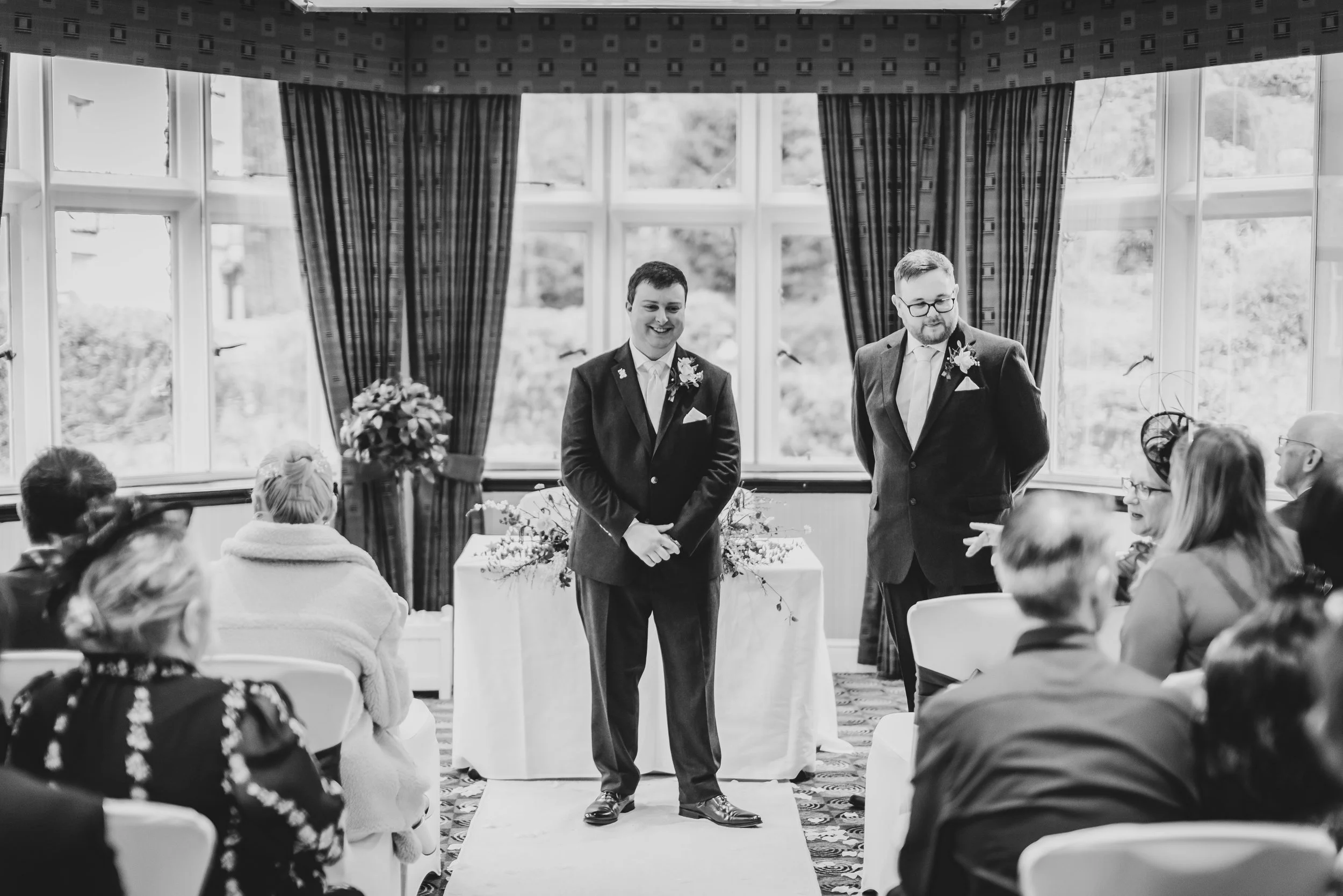 Two men in suits standing at the front of a small wedding ceremony in a brightly lit room with large windows and dark curtains, with guests seated facing them capture by Malvern Wedding Photographer