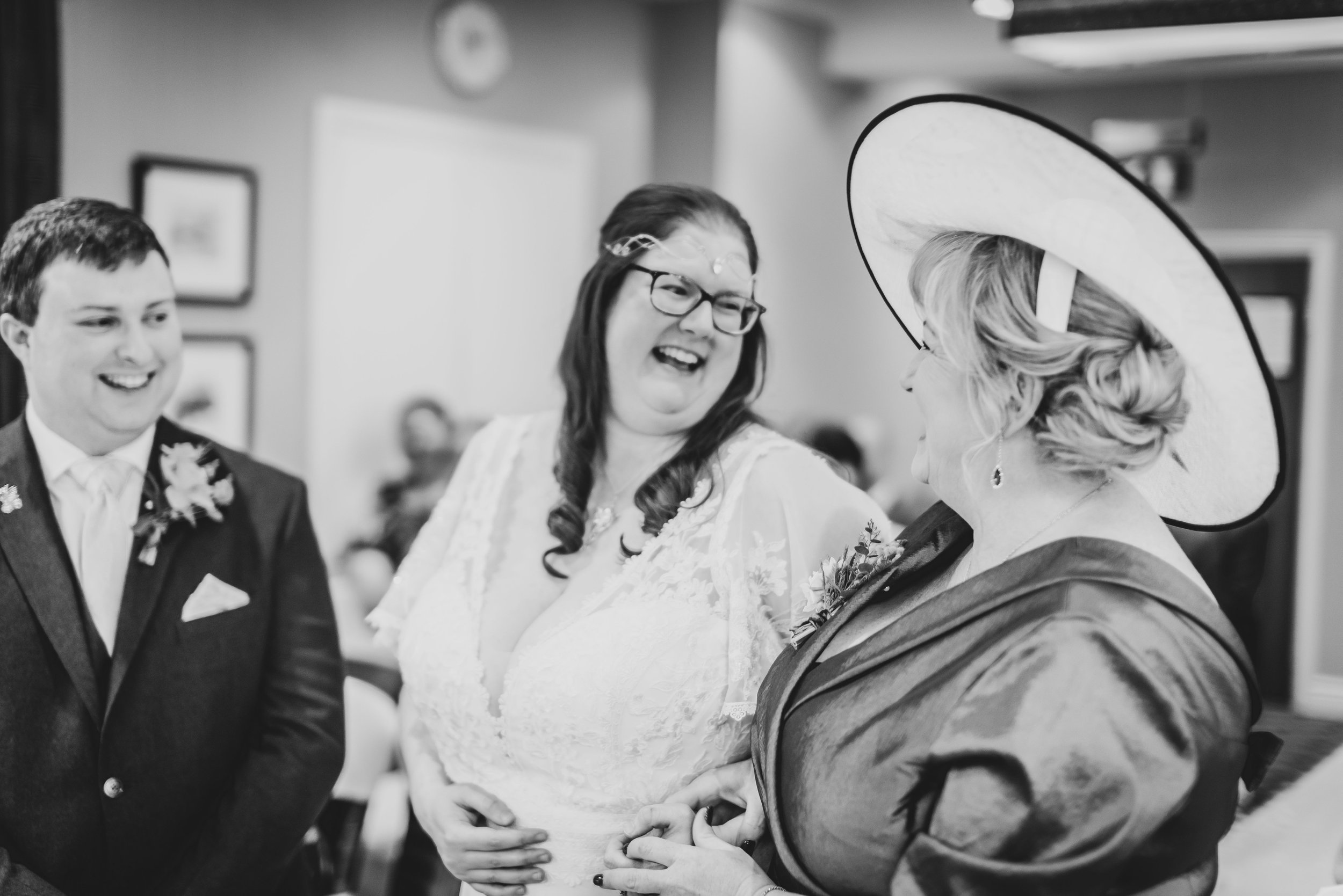 Black and white photo of three people at a wedding, smiling and holding hands. Mother of the Bride is wearing a large hat and glasses, the bride in the center has long dark hair and glasses, and the groom on the left is in a tuxedo.