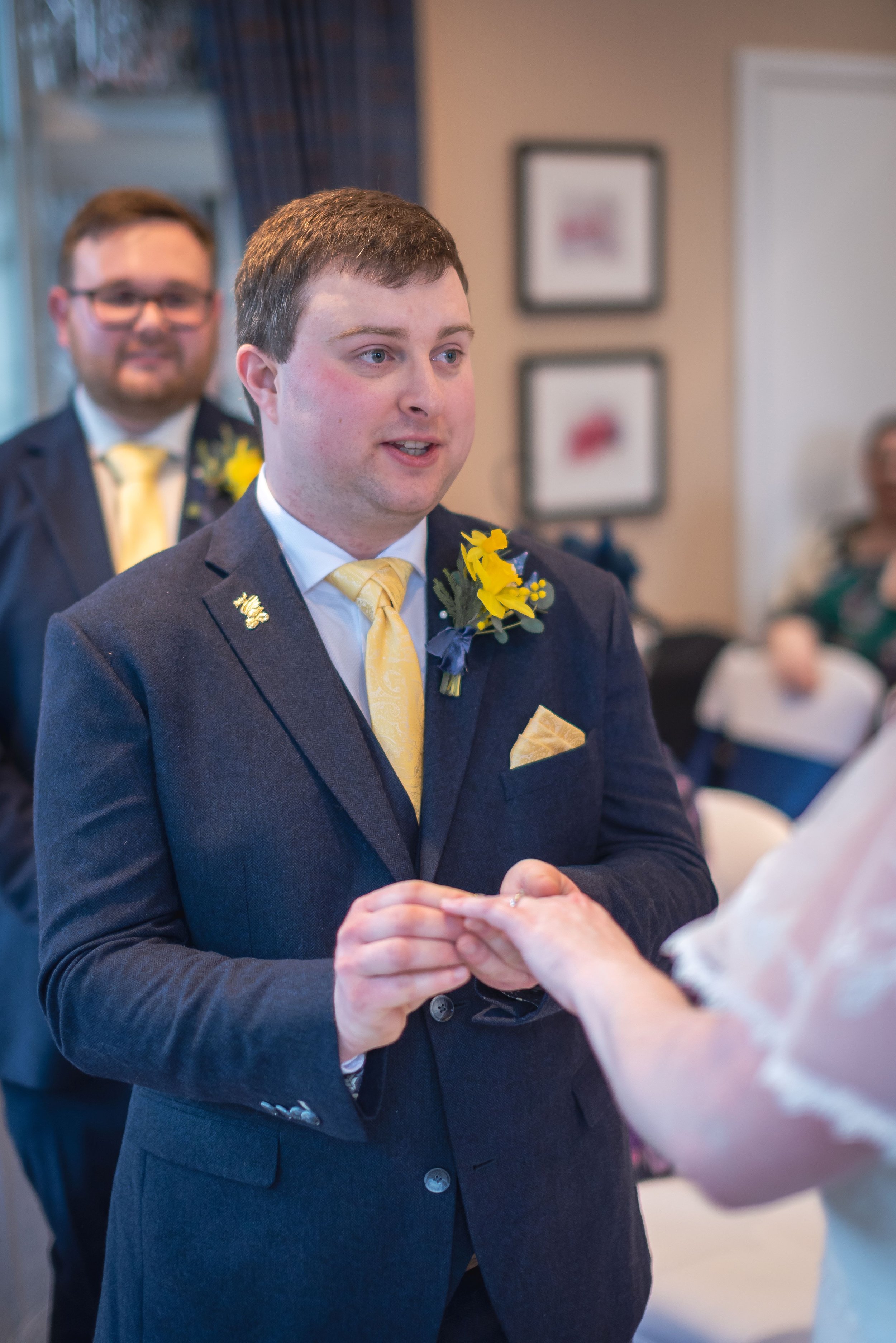 A groom exchanging rings during a wedding ceremony, with the officiant visible in the background during wedding at Abbey Hotel in Malvern shot by local wedding photographer.