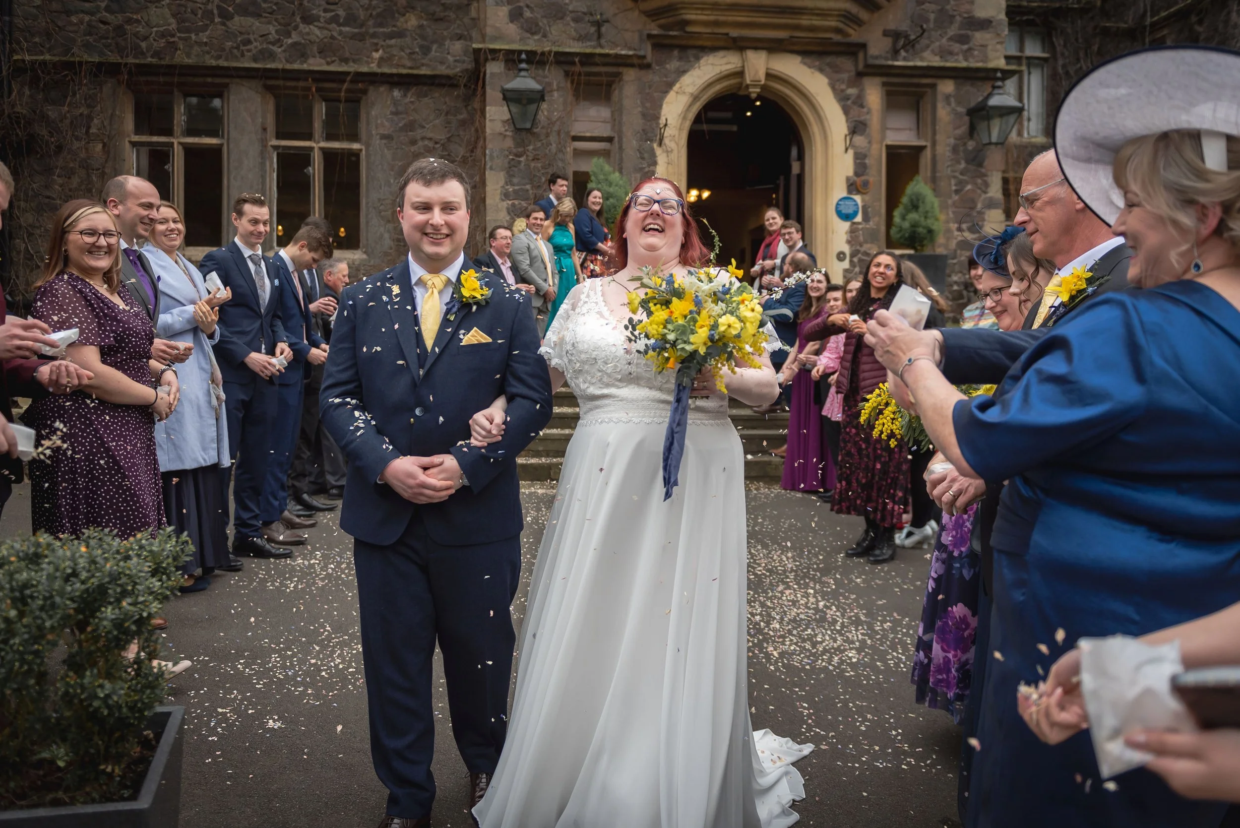 A bride and groom are walking out of a wedding reception at Abbey Hotel, smiling and throwing confetti. They are surrounded by happy guests outside a stone building. The bride is holding a bouquet of yellow flowers
