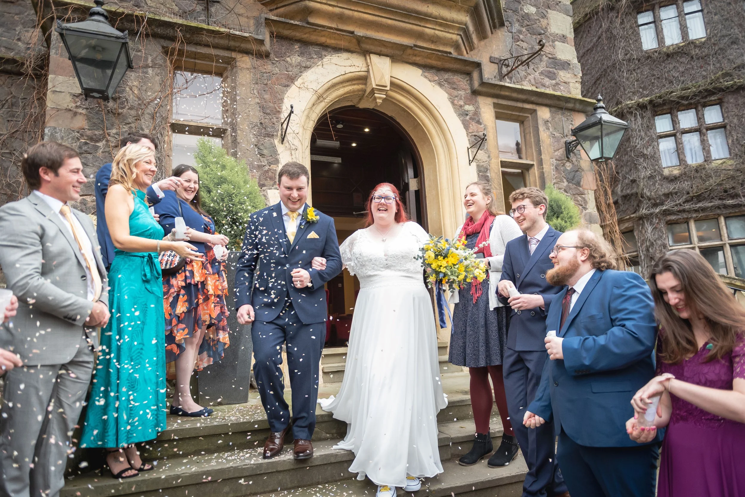 A group of people, including a bride in a white dress and a groom in a navy suit, celebrating on the steps outside a brick and stone building. The crowd is throwing confetti and smiling as they celebrate the wedding of the happy couple at Abbey Hotel