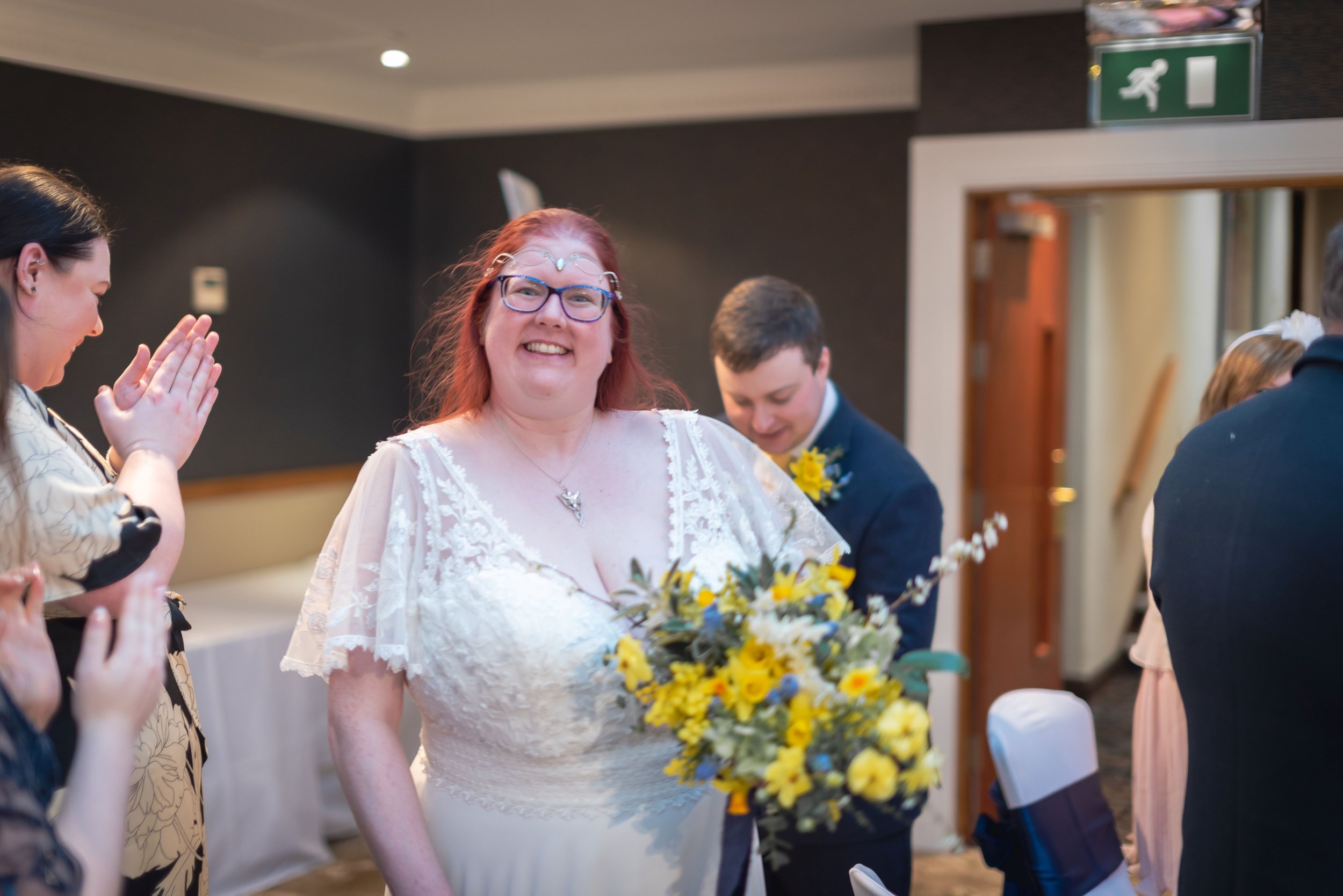 Photographer captures the moment the bride in a white wedding dress holding a bouquet of yellow flowers, smiling at a wedding reception surrounded by friends and family.
