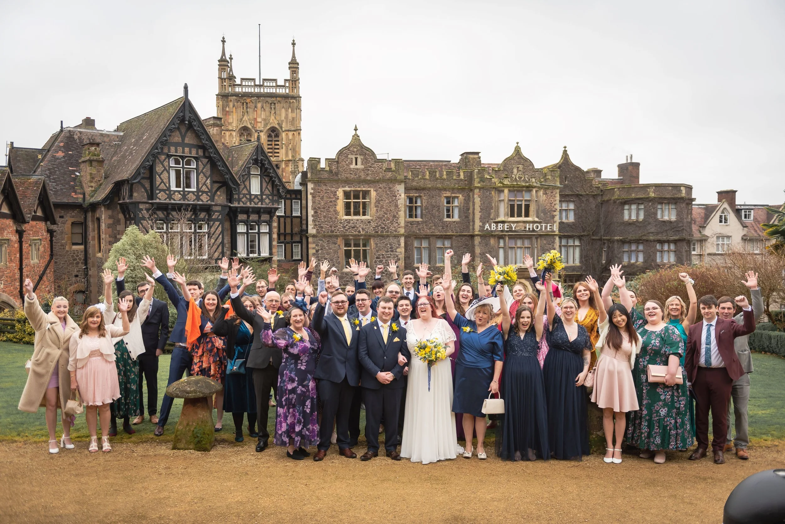 Gloucester wedding photographer coordinates group of people celebrating a wedding outside a historic building with a sign that reads 'Abbey Hotel'