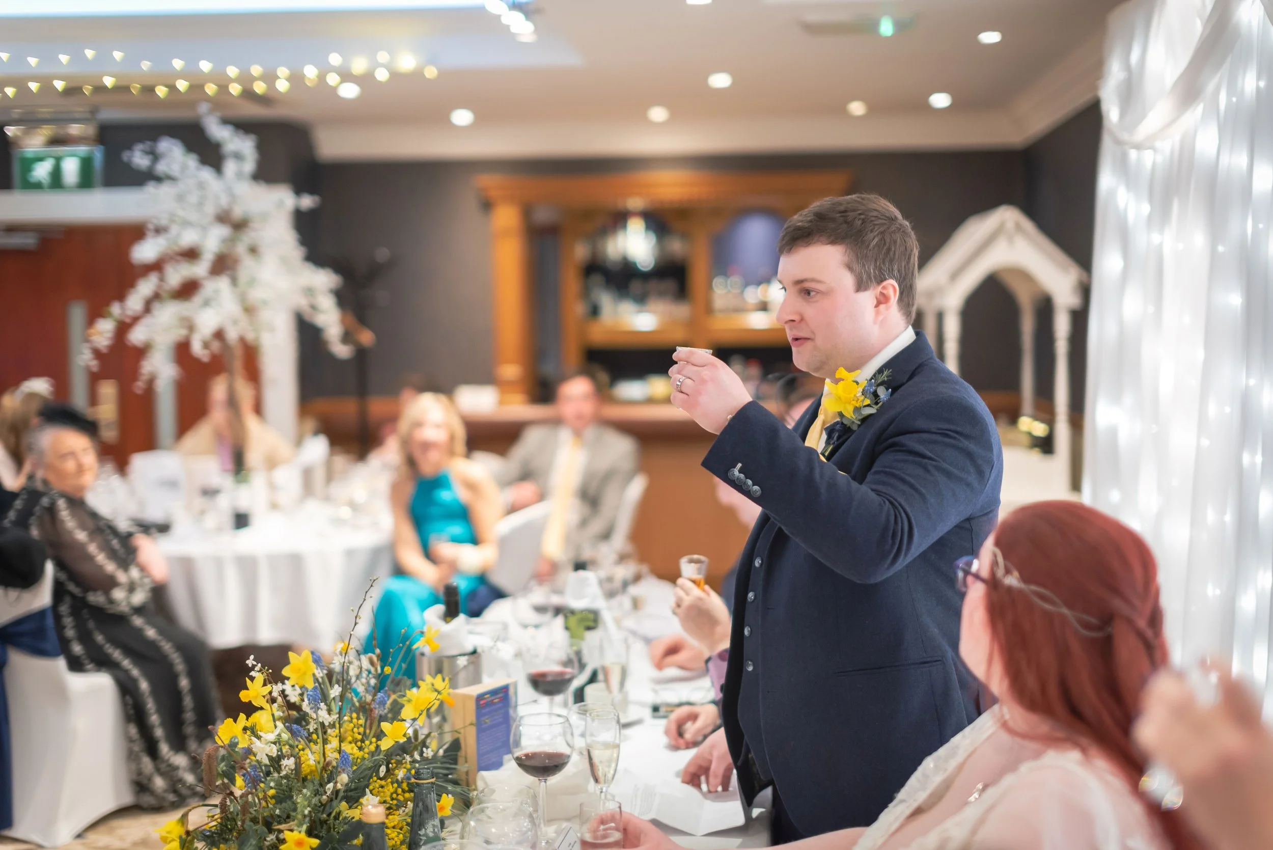 The groom in a navy suit with a yellow boutonniere giving a toast at a wedding reception, with guests seated at tables in the background decorated with flowers and white fairy lights in a moment captured by Gloucester Wedding Photographer