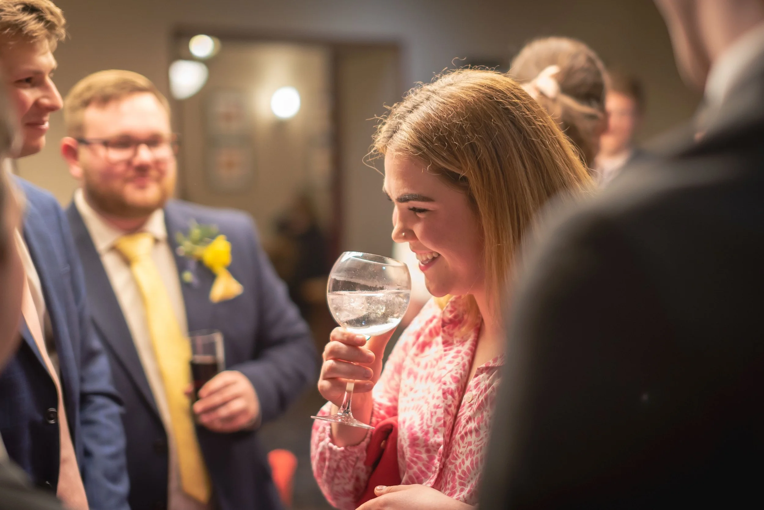 A group of people socializing at a party, with a woman smiling and holding a large glass of water. The scene is indoors with warm lighting.