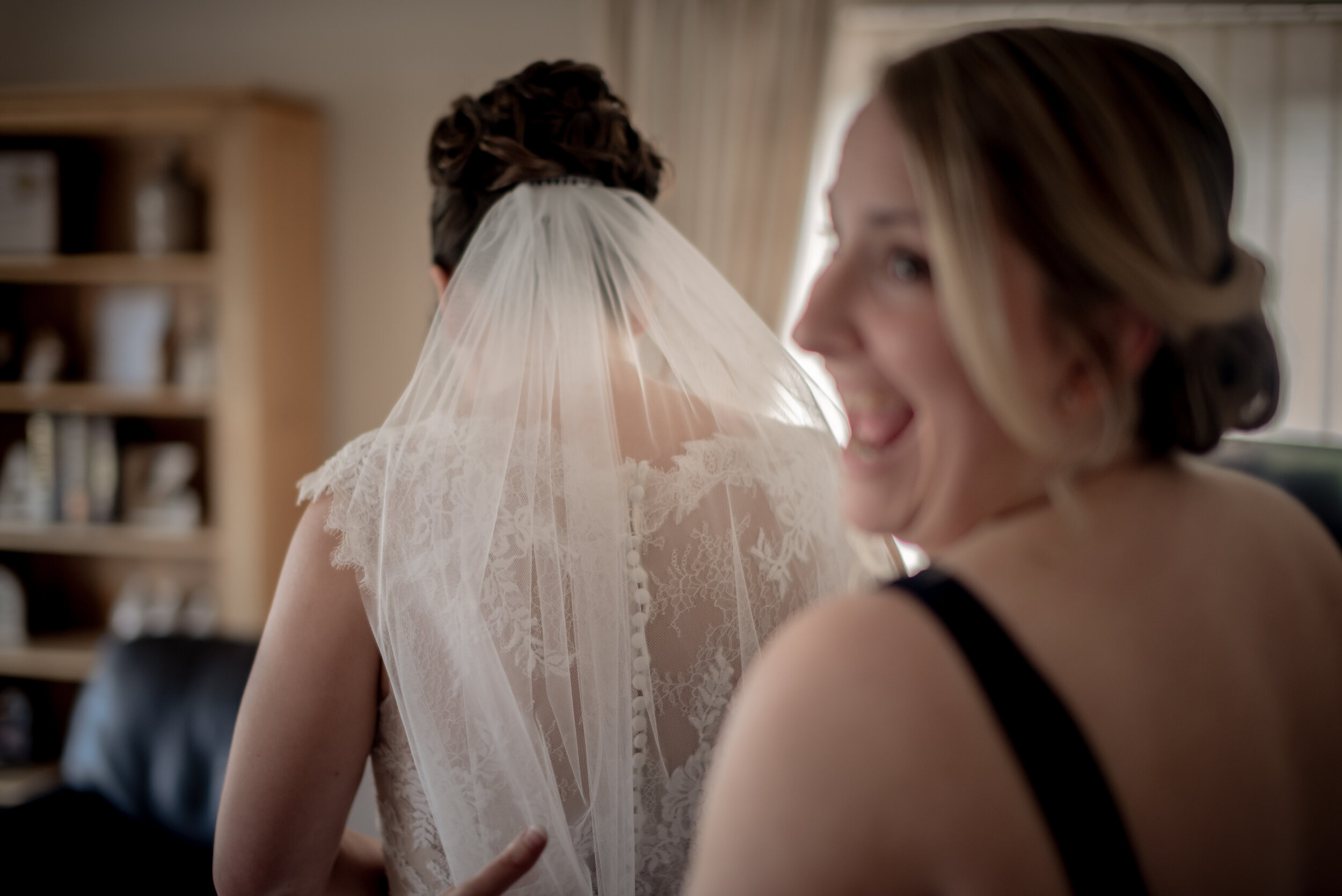 A bride in a wedding dress with a lace back and a veil, with a woman smiling and looking at her, in a softly lit indoor setting.