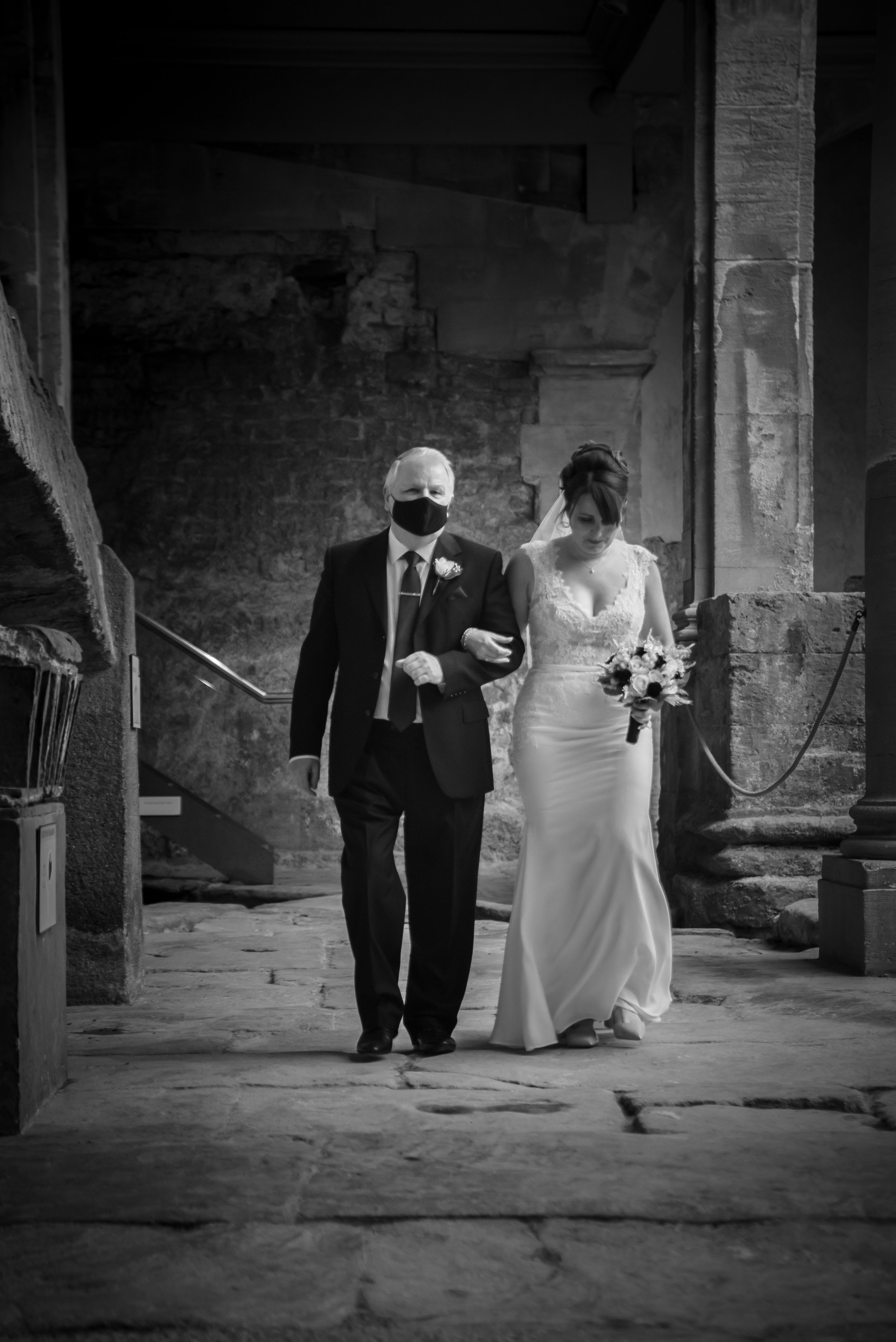 A bride walking with her father, who is wearing a mask, down the aisle inside at Roman Baths.
