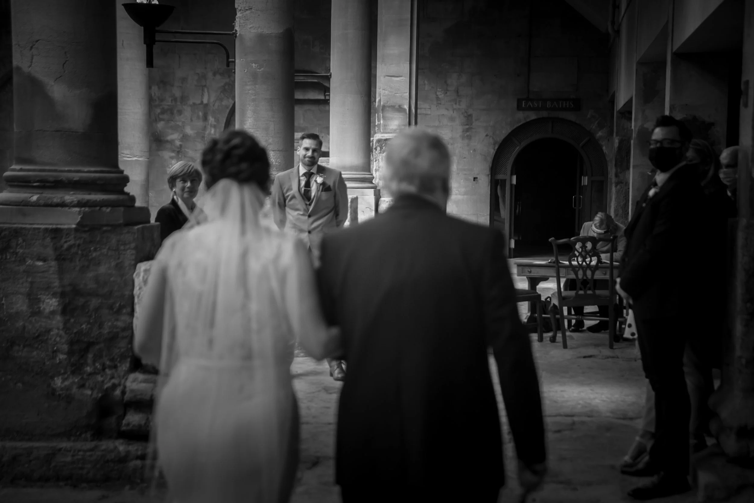 Black and white photo of a wedding ceremony inside a church, with a bride and groom facing an older man, and several guests standing nearby at Roman Baths.