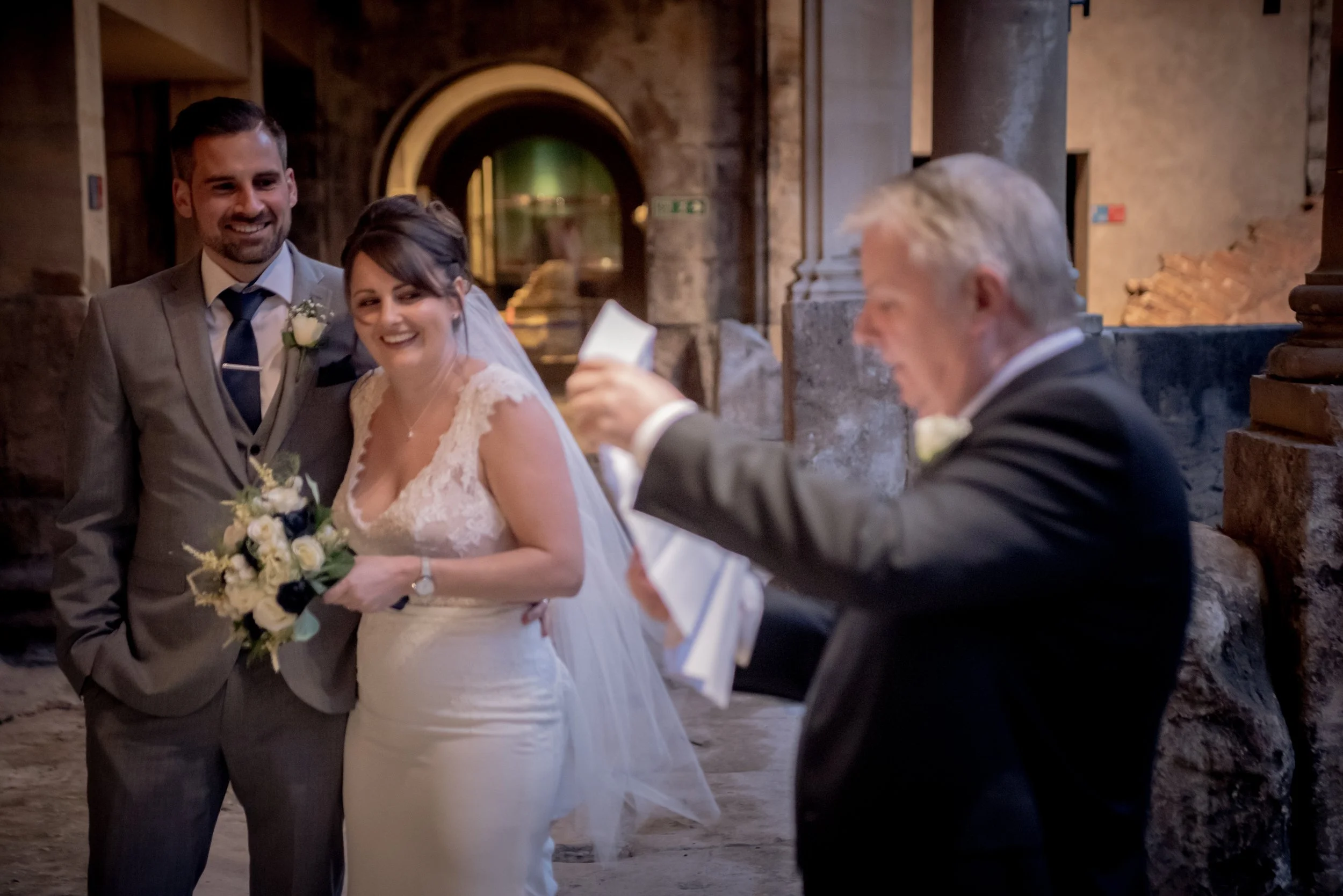 A wedding ceremony with a bride and groom smiling as they listen to an older man reading vows or a speech. The bride is holding a bouquet, and the setting appears to be in a historic or rustic venue with stone and brick background.