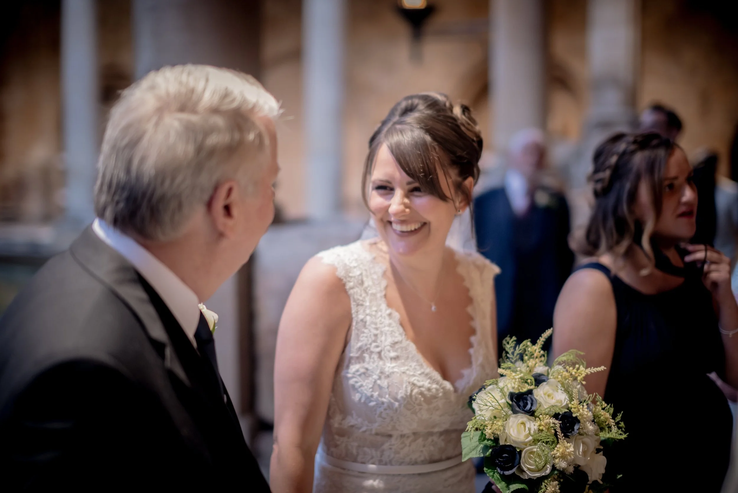 A bride smiling and holding a bouquet of white and dark flowers, standing beside a man in a suit at a wedding reception captured by Bath Wedding Photographer