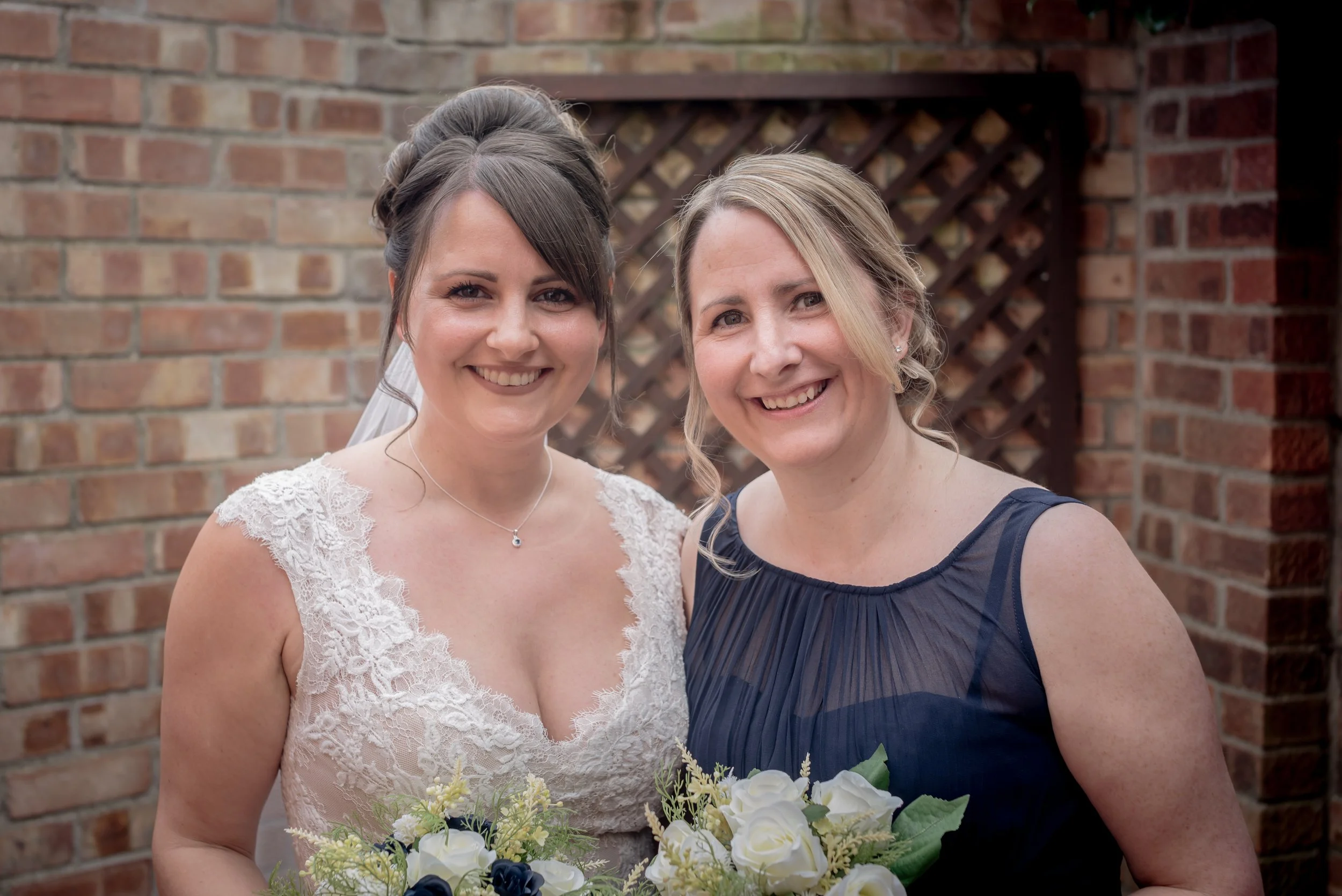 Two women smiling, one in a wedding dress holding a bouquet, and the other in a dark dress also holding a bouquet, standing outdoors in front of a brick wall at home address after bridal prep.