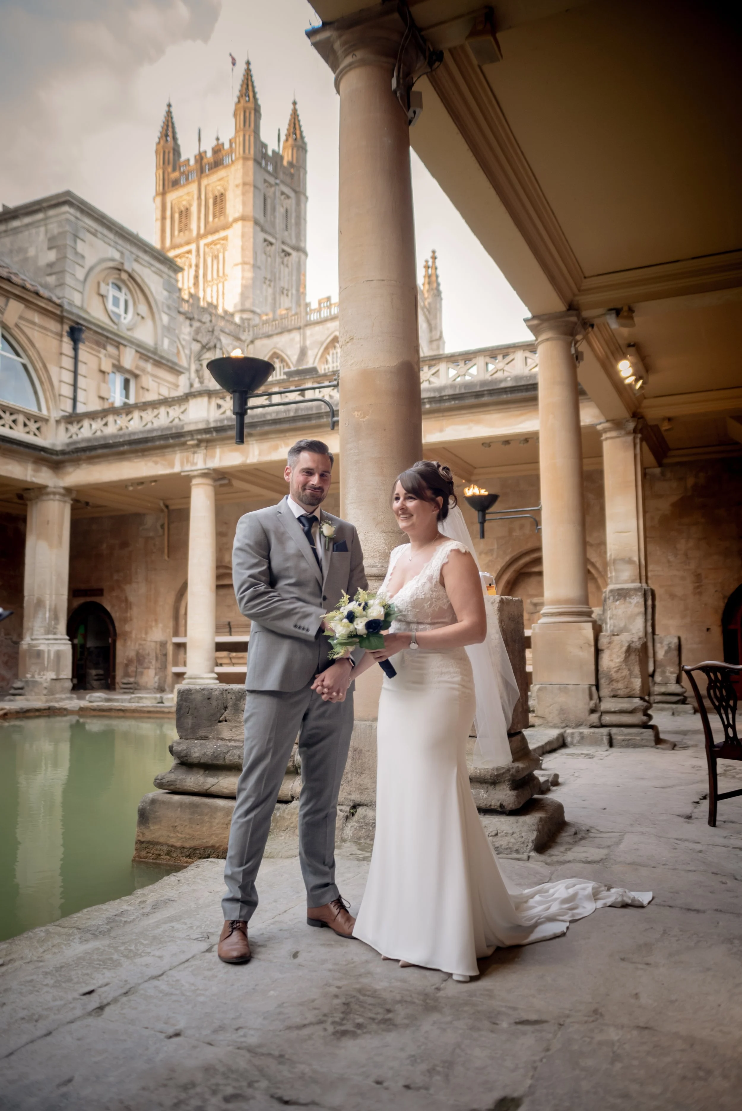 A bride and groom holding hands and smiling during their wedding, standing on stone pavement next to a pond with a historic building and church tower in the background, under a covered walkway with columns at Roman Baths