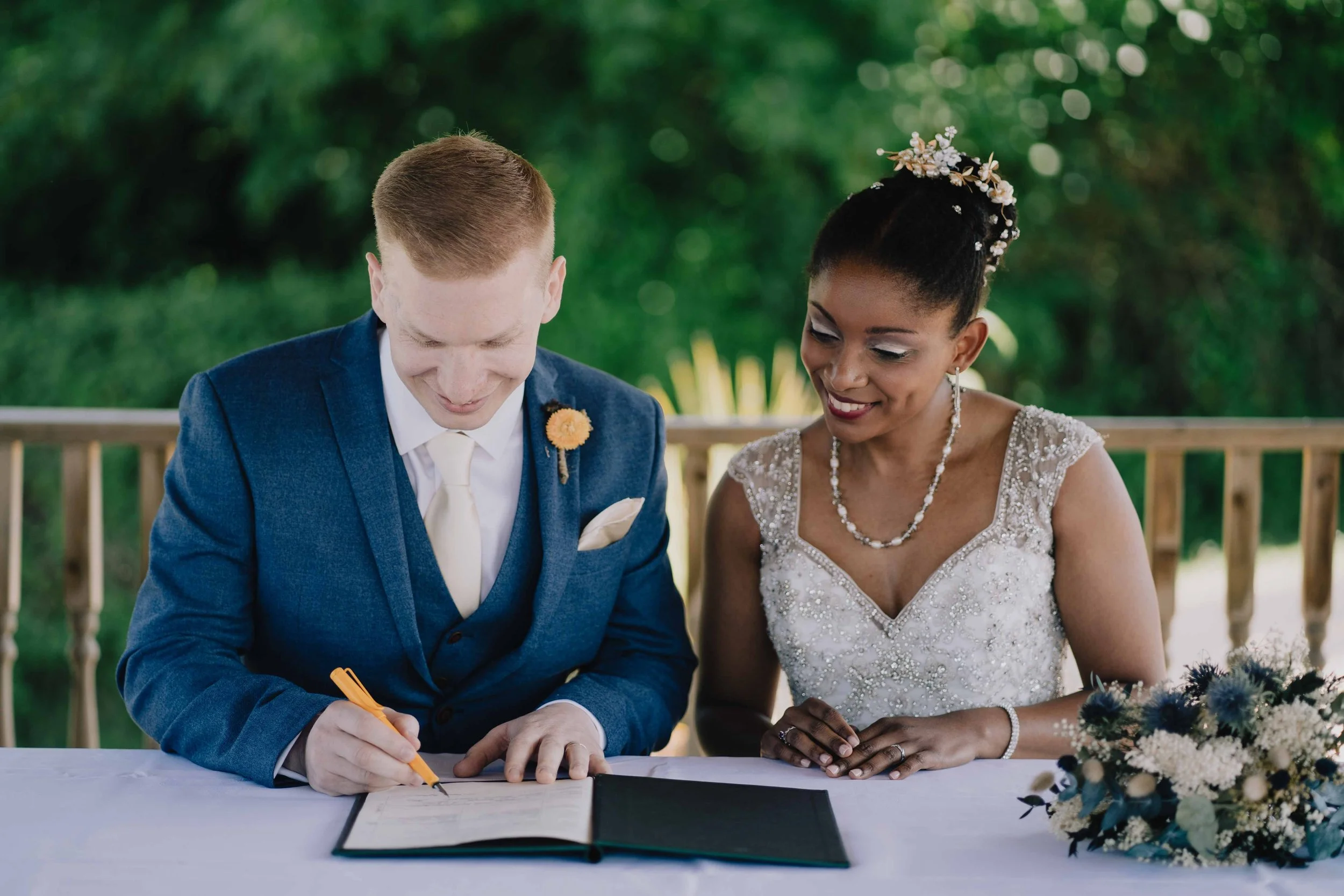 A bride and groom signing a marriage certificate at their wedding reception outdoors, with a bouquet of flowers on the table at Riverside near Bristol