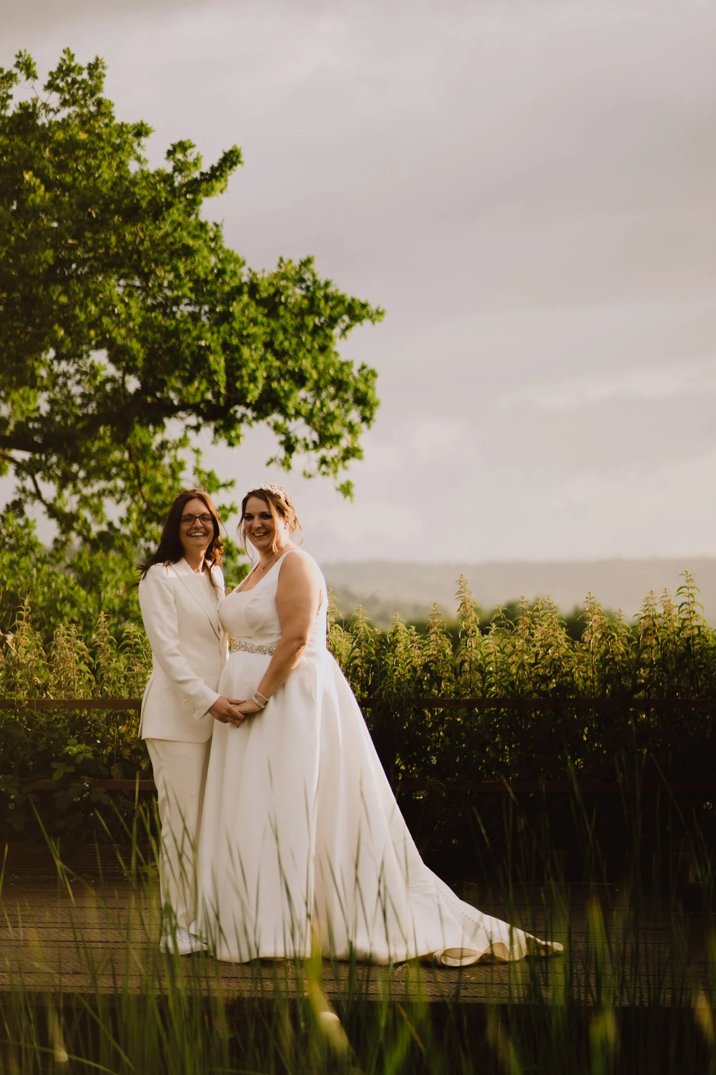 Two women in wedding dresses holding hands and smiling outdoors near a green tree and tall grass, on a cloudy day.