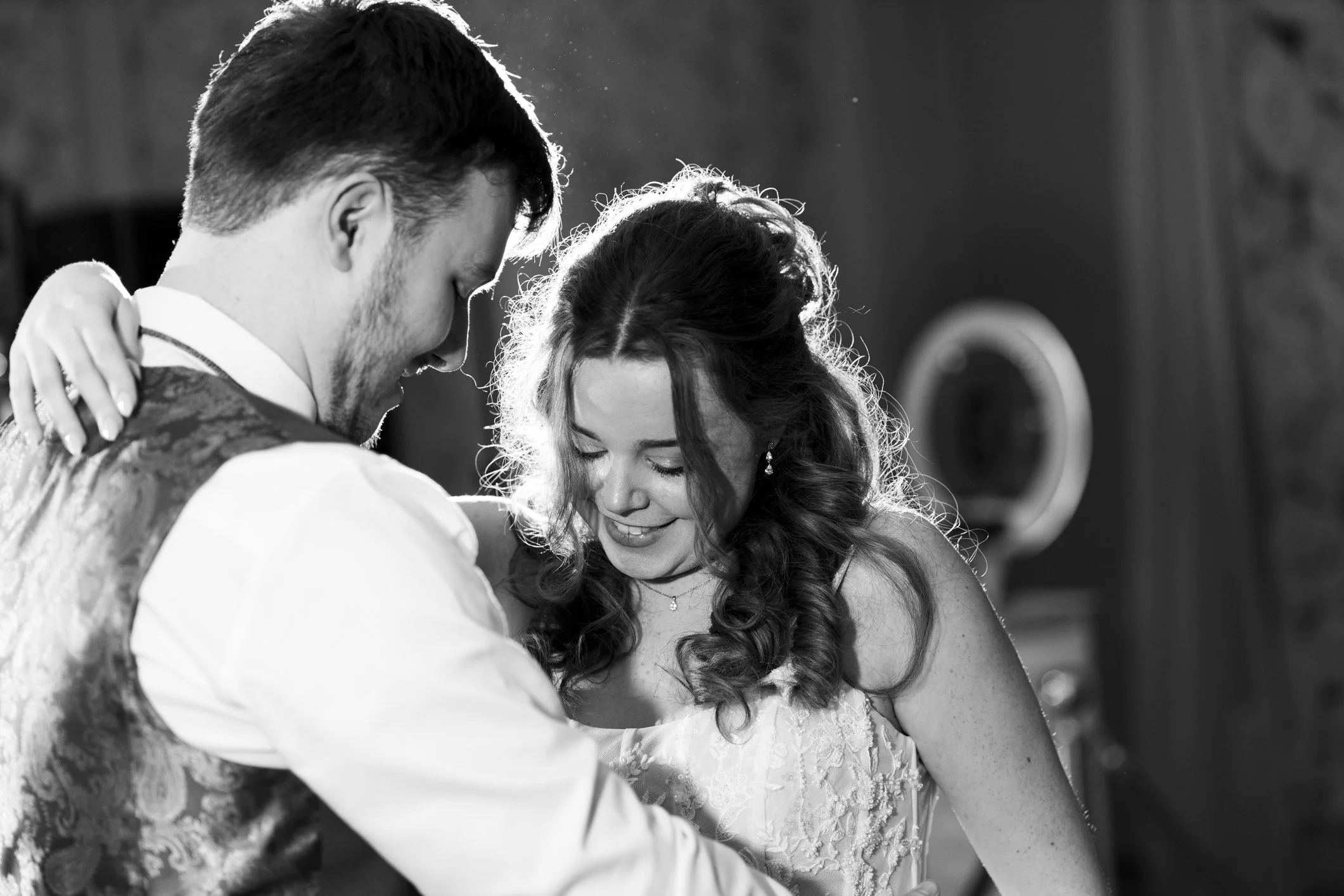 A black and white photo of a newlywed couple sharing a dance, smiling and embracing each other closely.