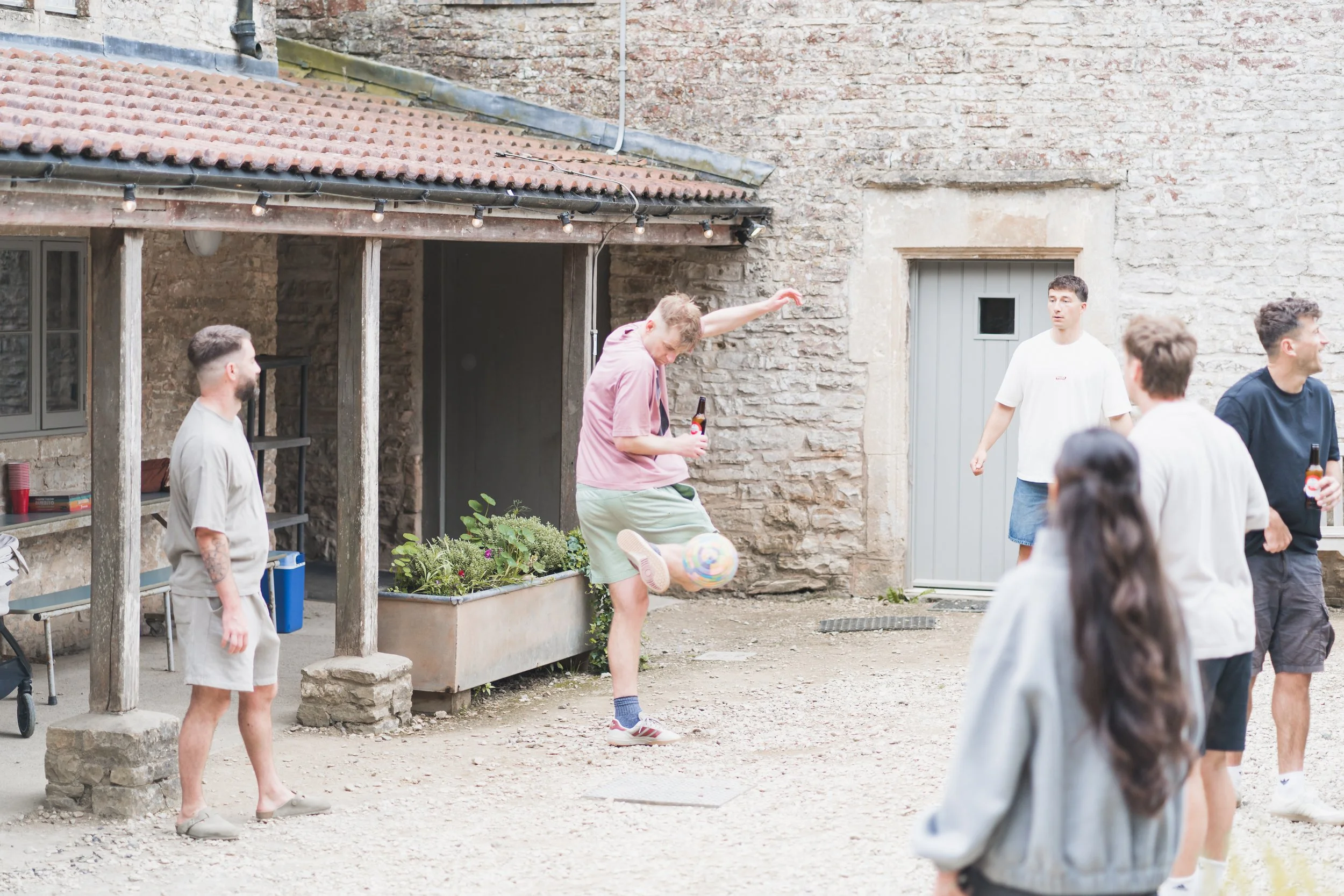 People playing soccer outside on a gravel area next to a stone building with a red tiled roof at Folly Farm. One person is mid-kick with a soccer ball, while others watch or stand nearby.