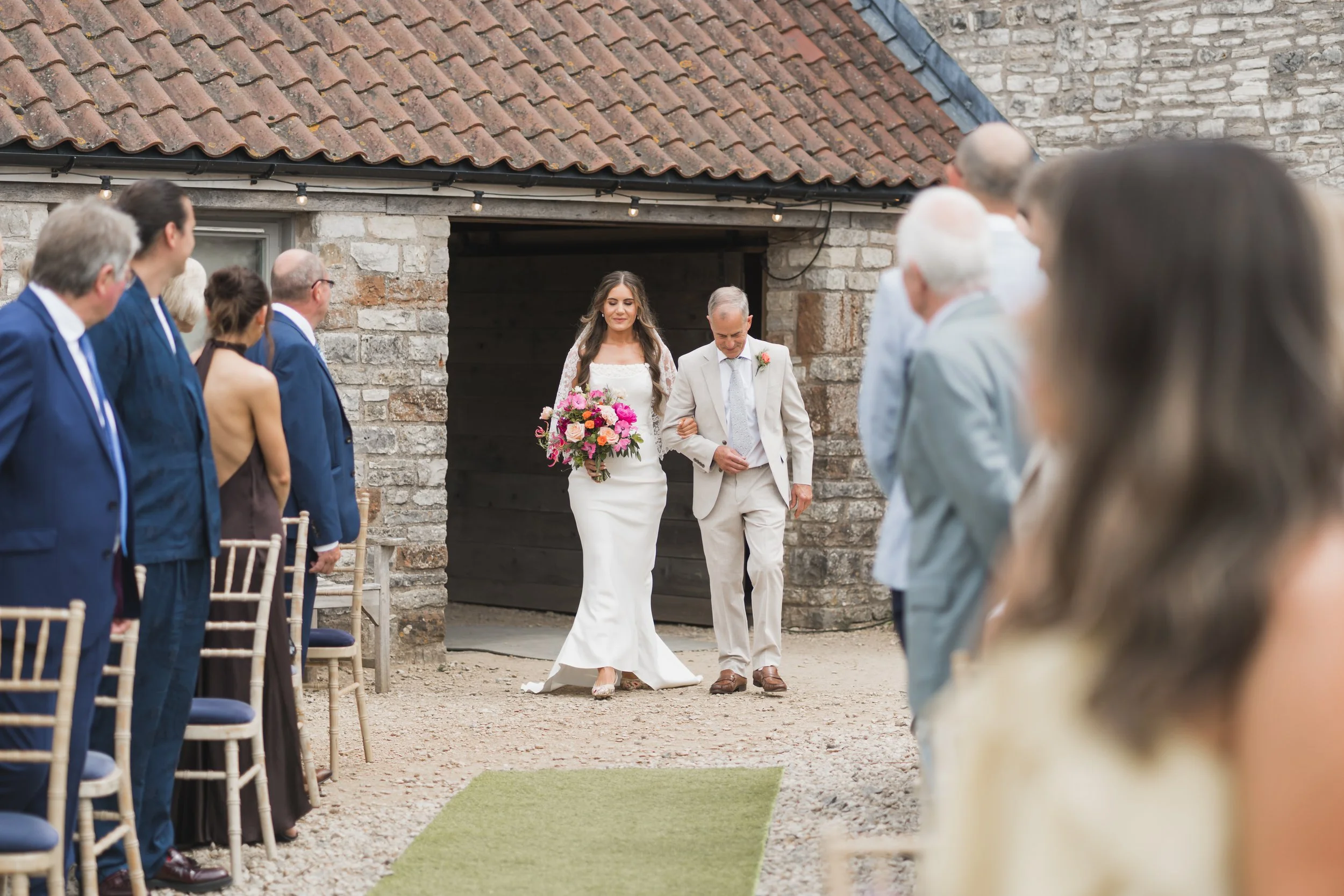 A bride walking down the aisle with her father during an outdoor wedding ceremony held at Folly Farm, with guests seated on either side.