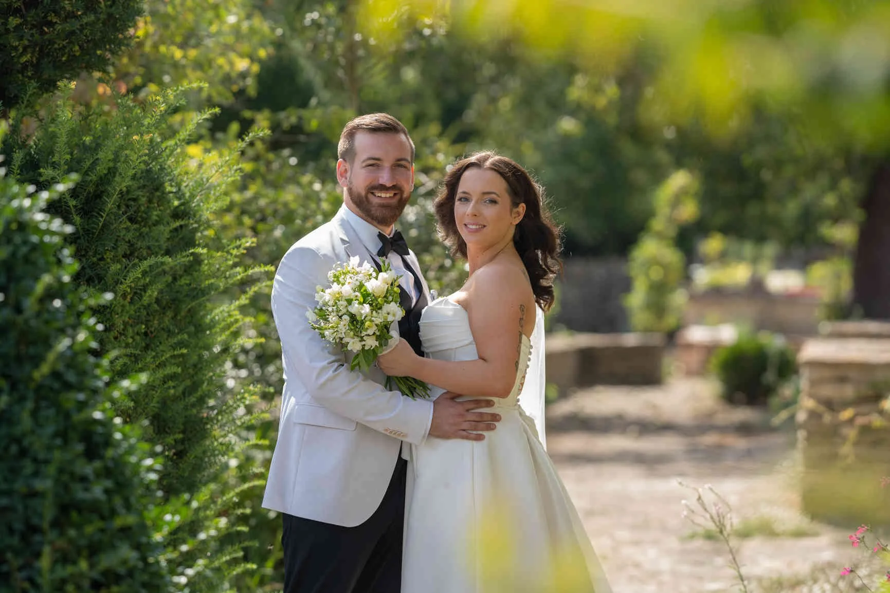 A newlywed couple in wedding attire standing outdoors on a sunny day at Oxleaze Barn. The groom is wearing a white tuxedo jacket with a black bow tie, and the bride is in a strapless white gown holding a bouquet of white flowers.