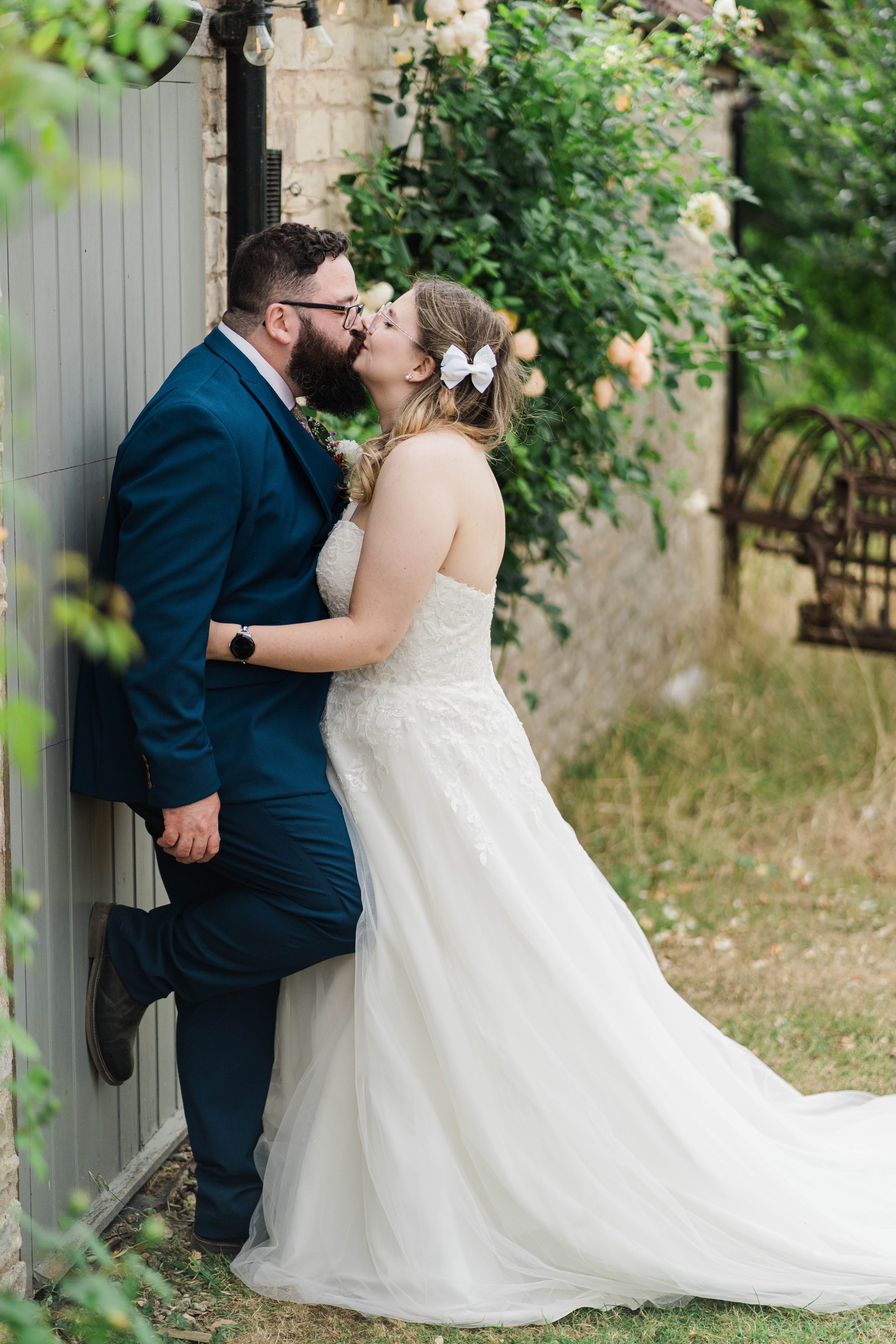 A bride and groom kiss outdoors near a brick wall and greenery, the bride in a white strapless wedding gown with a bow in her hair, and the groom in a dark blue suit with glasses, holding her around the waist.