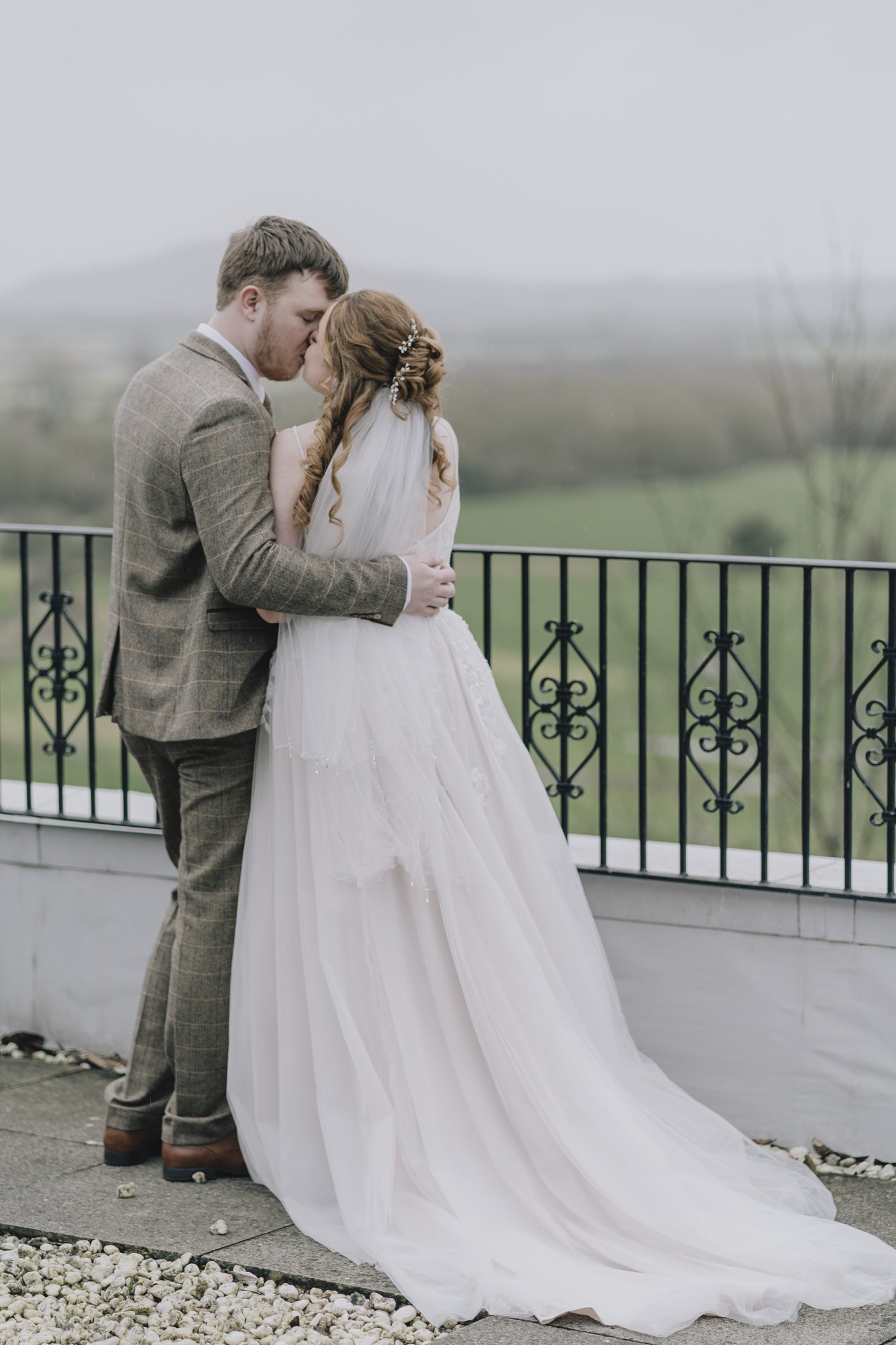 A bride and groom share a kiss on a balcony, with the bride in a white wedding dress and the groom in a brown plaid suit. The scene is outdoors with a scenic, foggy mendips in the background.