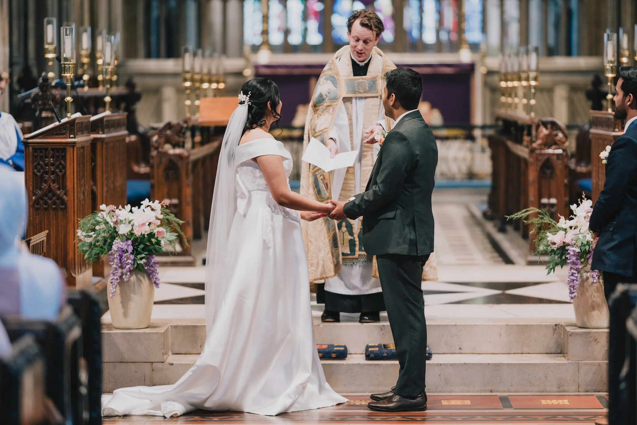 A wedding ceremony taking place in a Bristol church, with a bride and groom holding hands and facing each other, a priest officiating, and guests seated nearby.