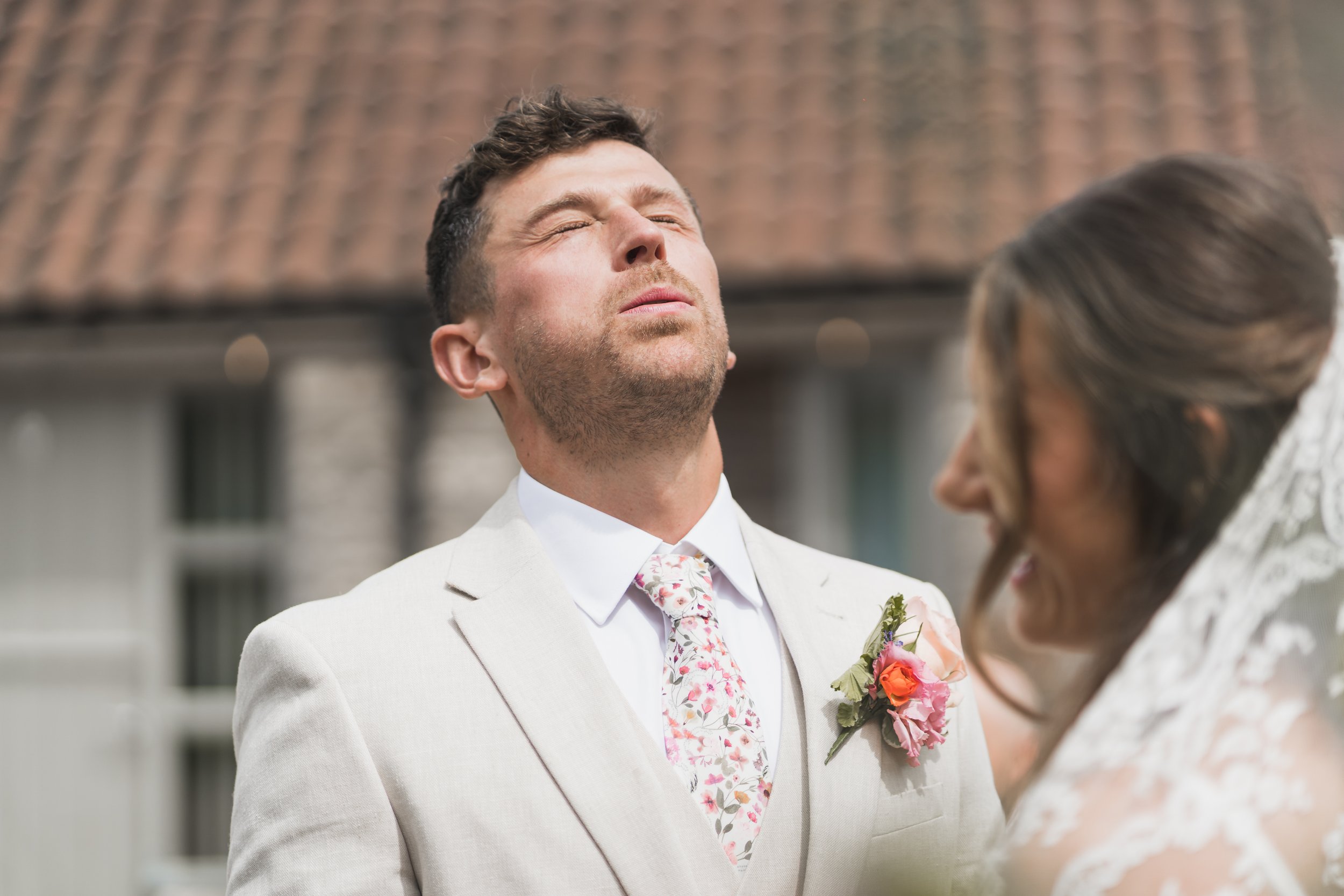 A groom with brown hair and a closed mouth, wearing a light-colored suit with a floral tie, standing outside with a blurred tiled roof in the background.