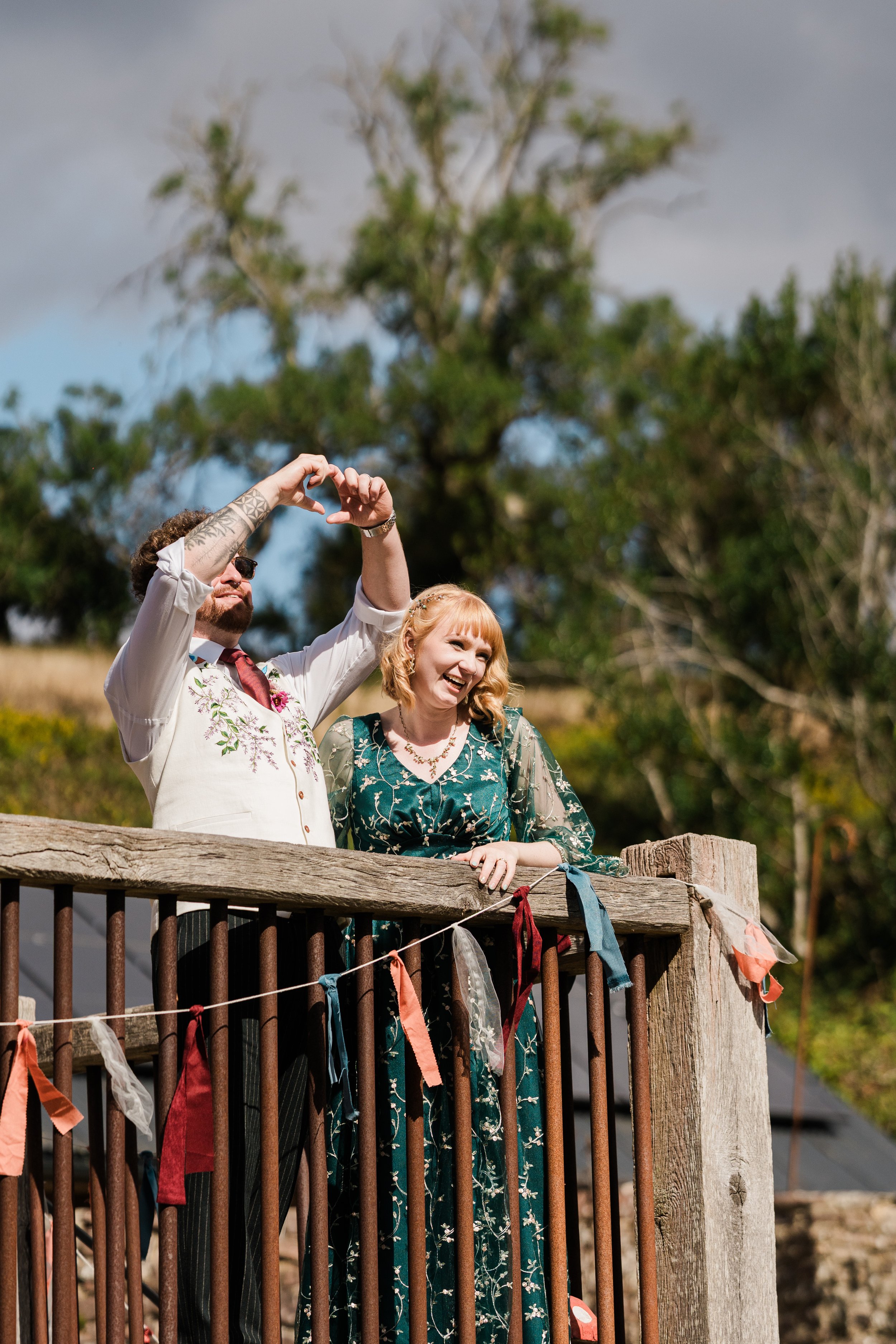 A couple stands on a wooden fence, taking a photo together during a outdoor event. The man has tattoos and is wearing sunglasses, a white shirt with embroidered flowers, and a maroon tie. The woman has blonde curly hair and is wearing a green floral dress with a sheer overlay. Ribbons and fabric strips are tied to the fence, and trees with green foliage are in the background under a cloudy sky.