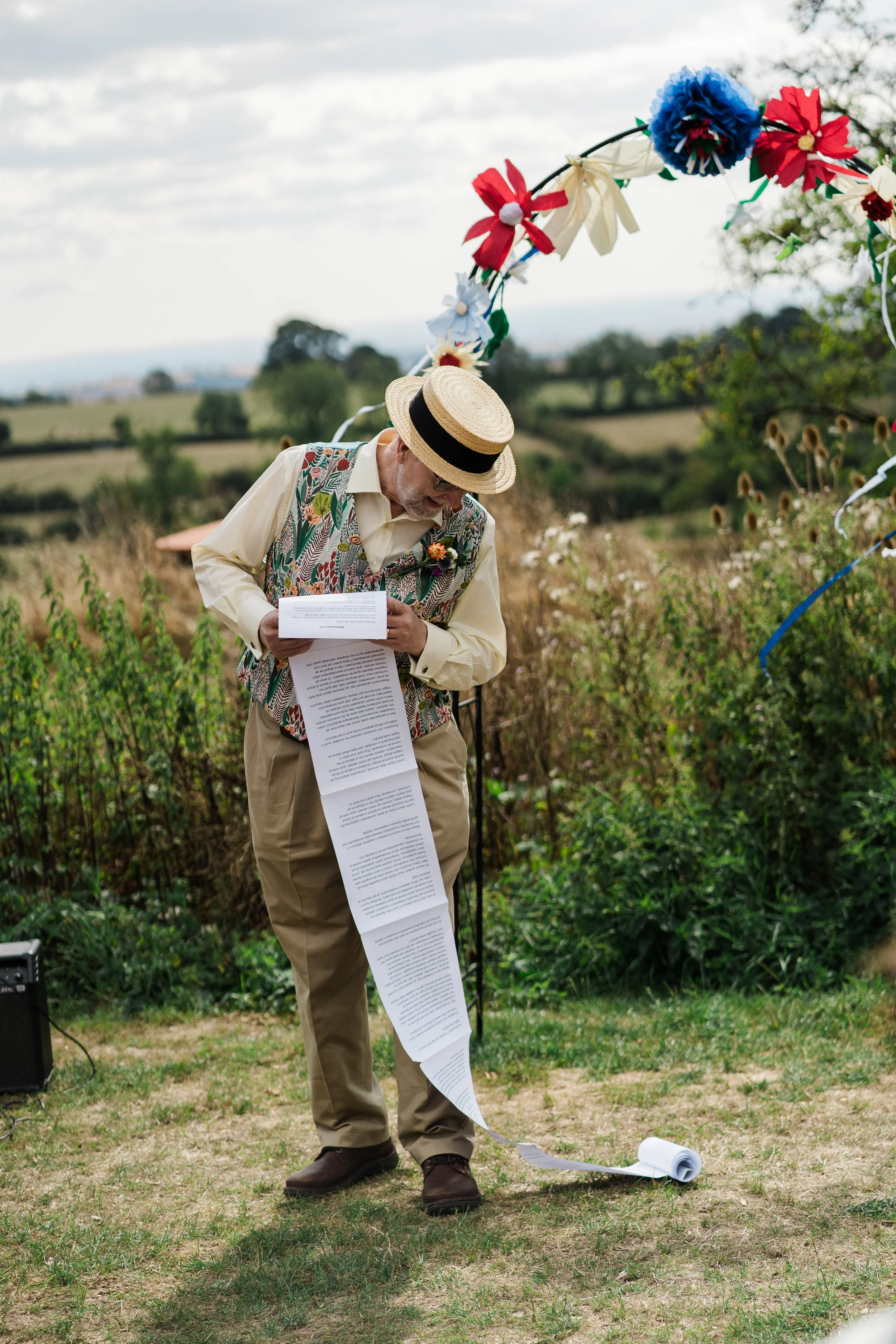 A man in a straw hat and colorful vest stands outdoors, holding a long sheet of paper, with a decorative floral arch behind him.